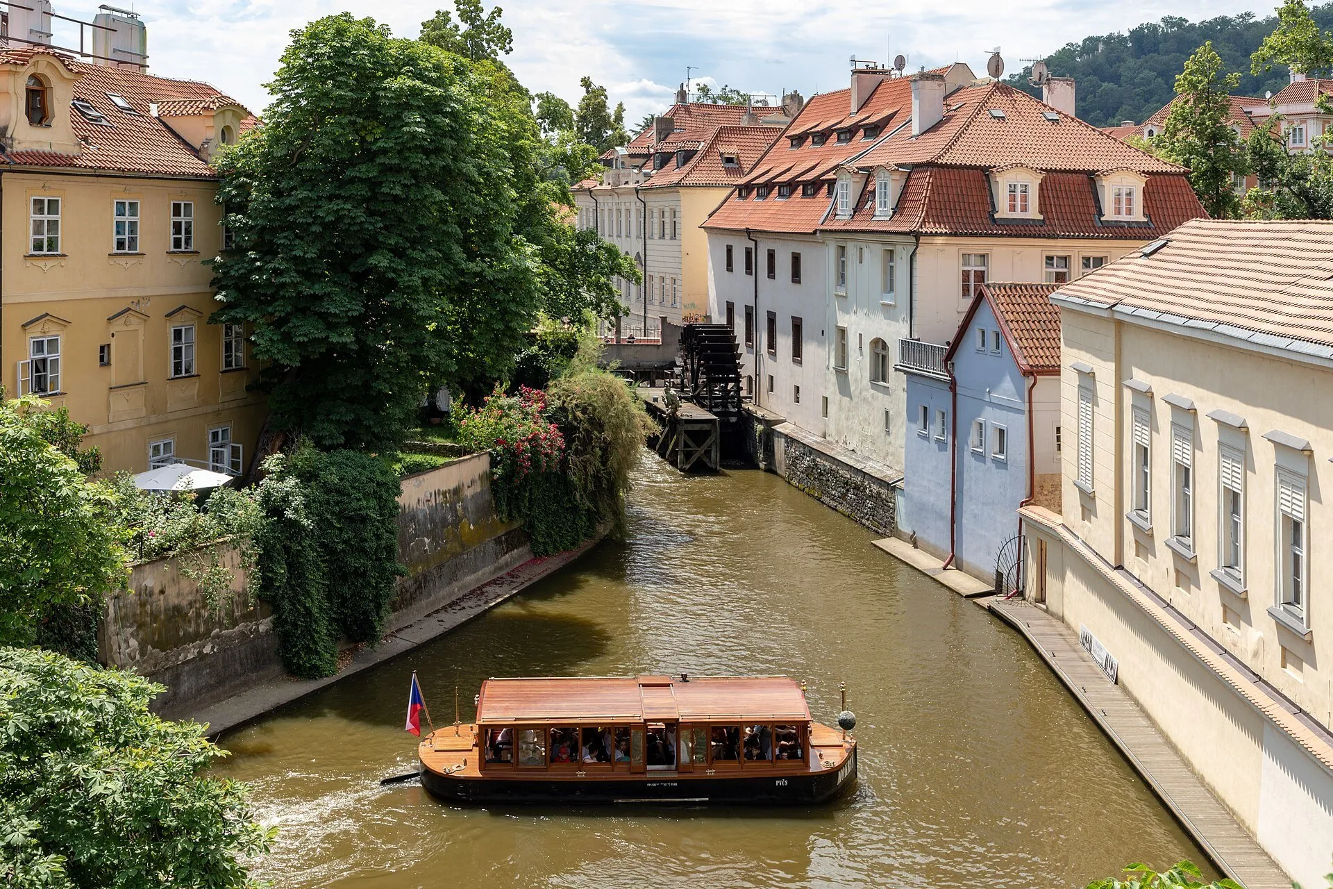 Canal Čertovka en Kampa, con rueda de molino, casas de tejados rojos, árboles y un barco de paseo.