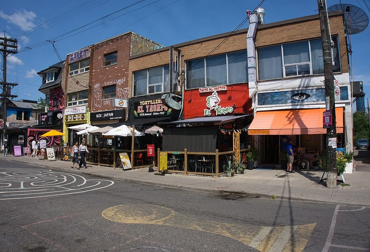 Lojas e restaurantes coloridos em Kensington Market, com toldos, murais e alguns pedestres sob céu azul.