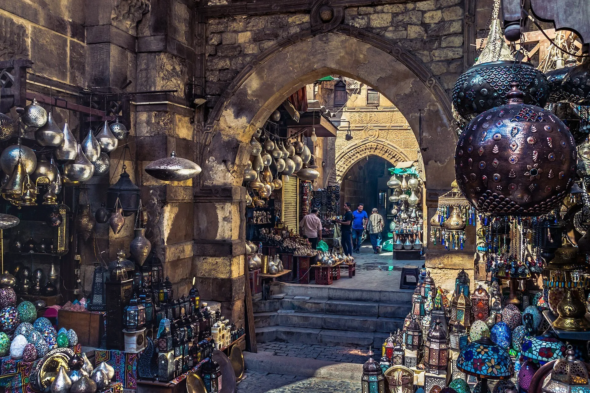 Passagem com arcos e escadas em Khan el-Khalili, ladeada por lojas de luminárias e objetos de metal.