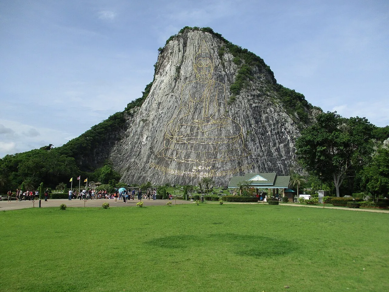 Buda dourado gravado na encosta rochosa de Khao Chi Chan, com gramado e pequenos visitantes à frente.