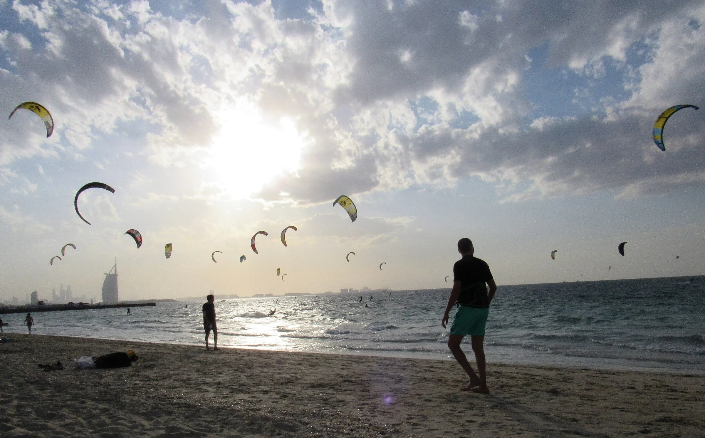 Dozens of kites in a cloudy sky over the sea at Kite Beach, with the Burj Al Arab silhouette behind and bathers on the sand.