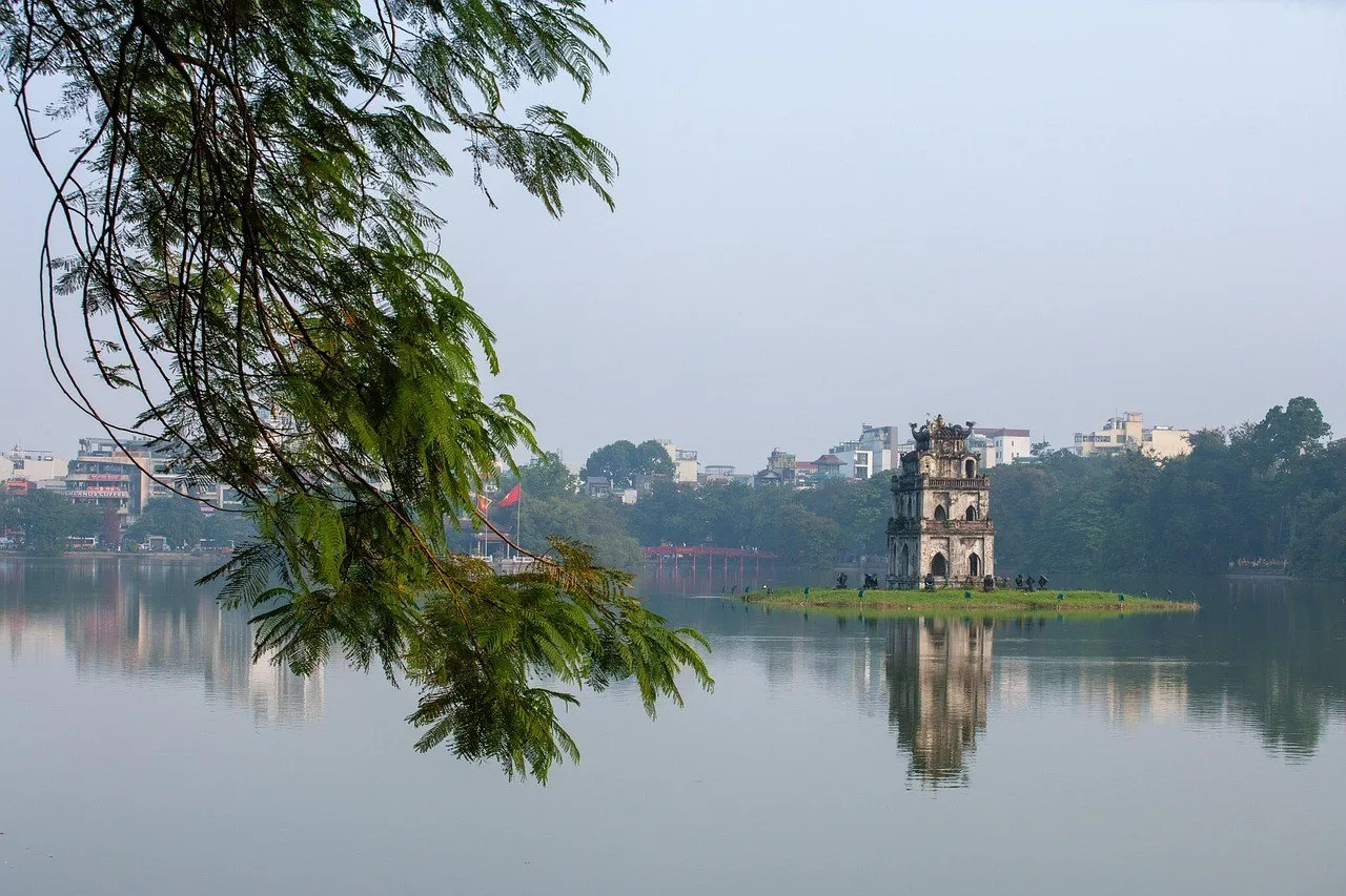Torre da Tartaruga refletida no lago Hoan Kiem, com galhos em primeiro plano e prédios ao fundo.