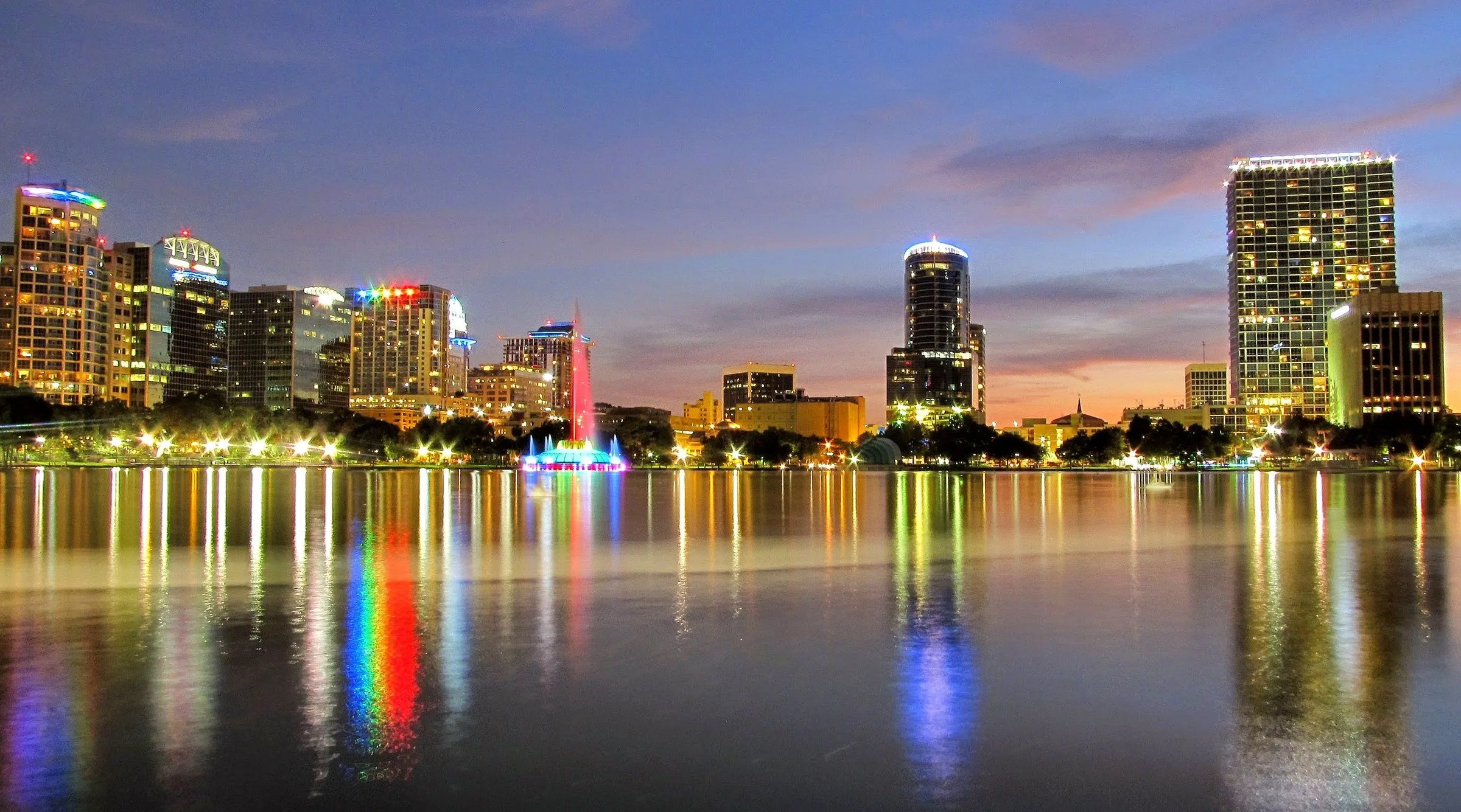 Fonte iluminada e prédios de Downtown Orlando refletidos no Lake Eola ao anoitecer.