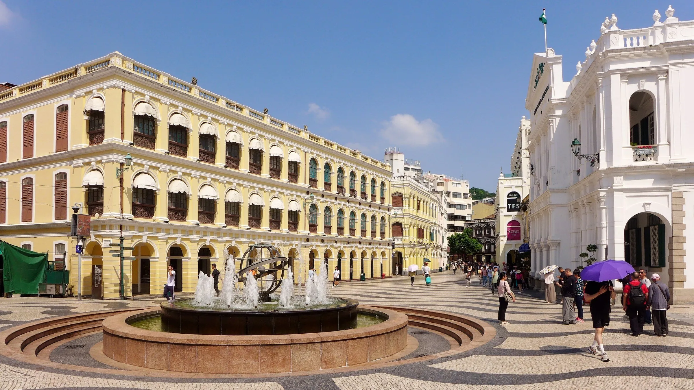 Plaza con empedrado ondulado en blanco y negro, fuente al centro y edificios históricos amarillos y blancos bajo cielo azul.