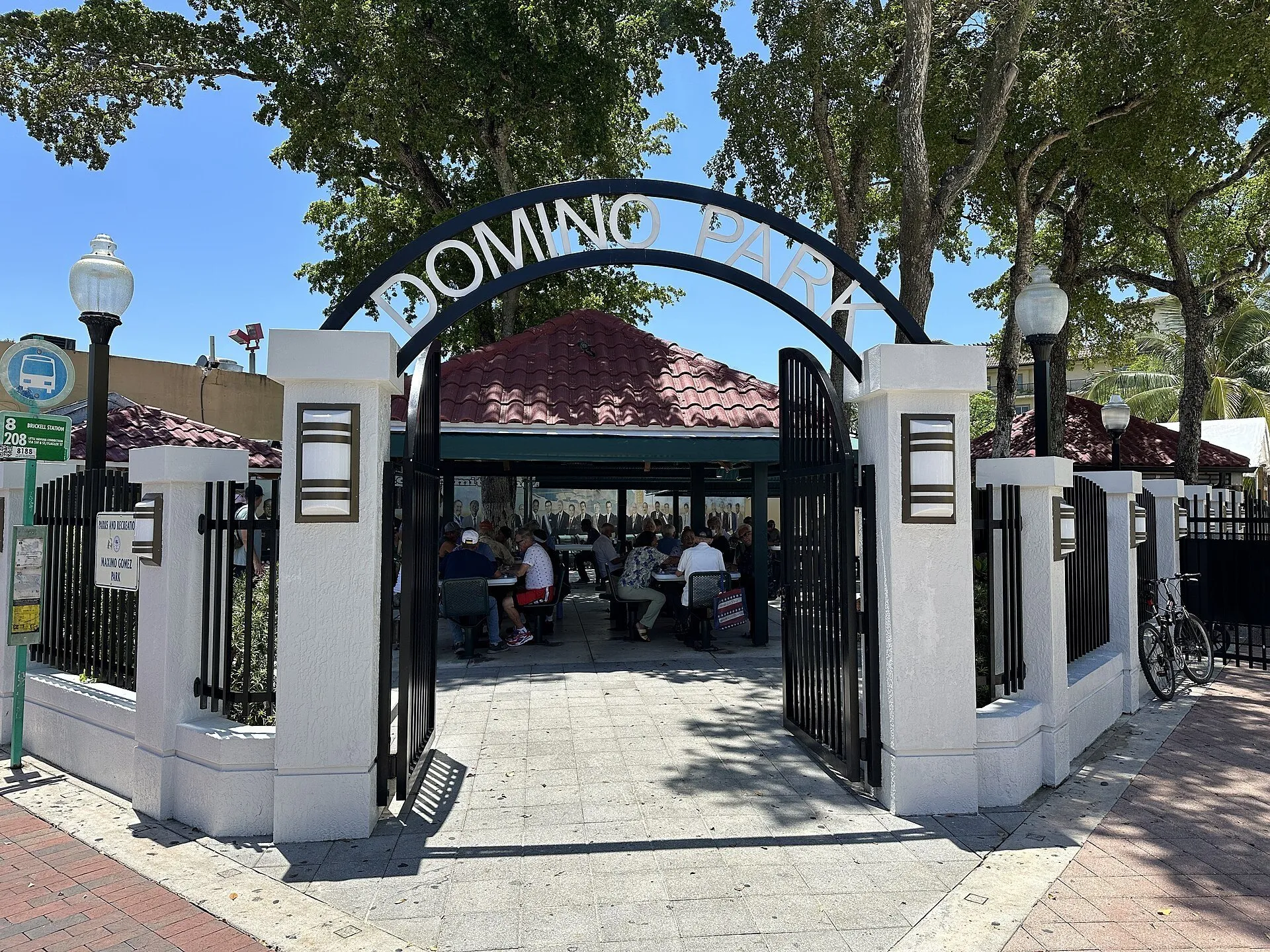 Entrada branca do Domino Park em Little Havana, com arco de letras, árvores e jogadores ao fundo.