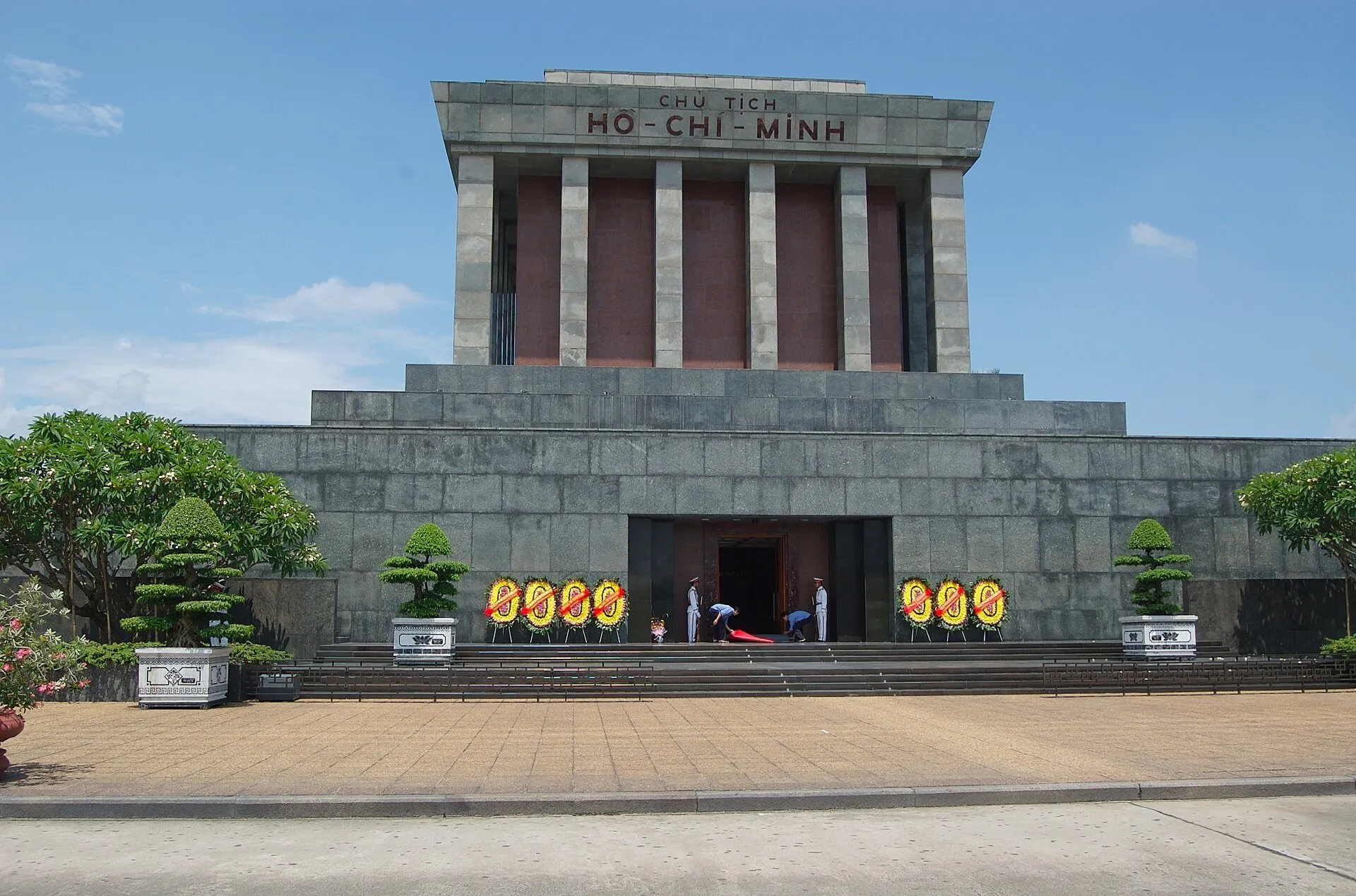 Fachada frontal de pedra do Mausoléu Ho Chi Minh, com inscrição, jardins em vasos e céu azul.