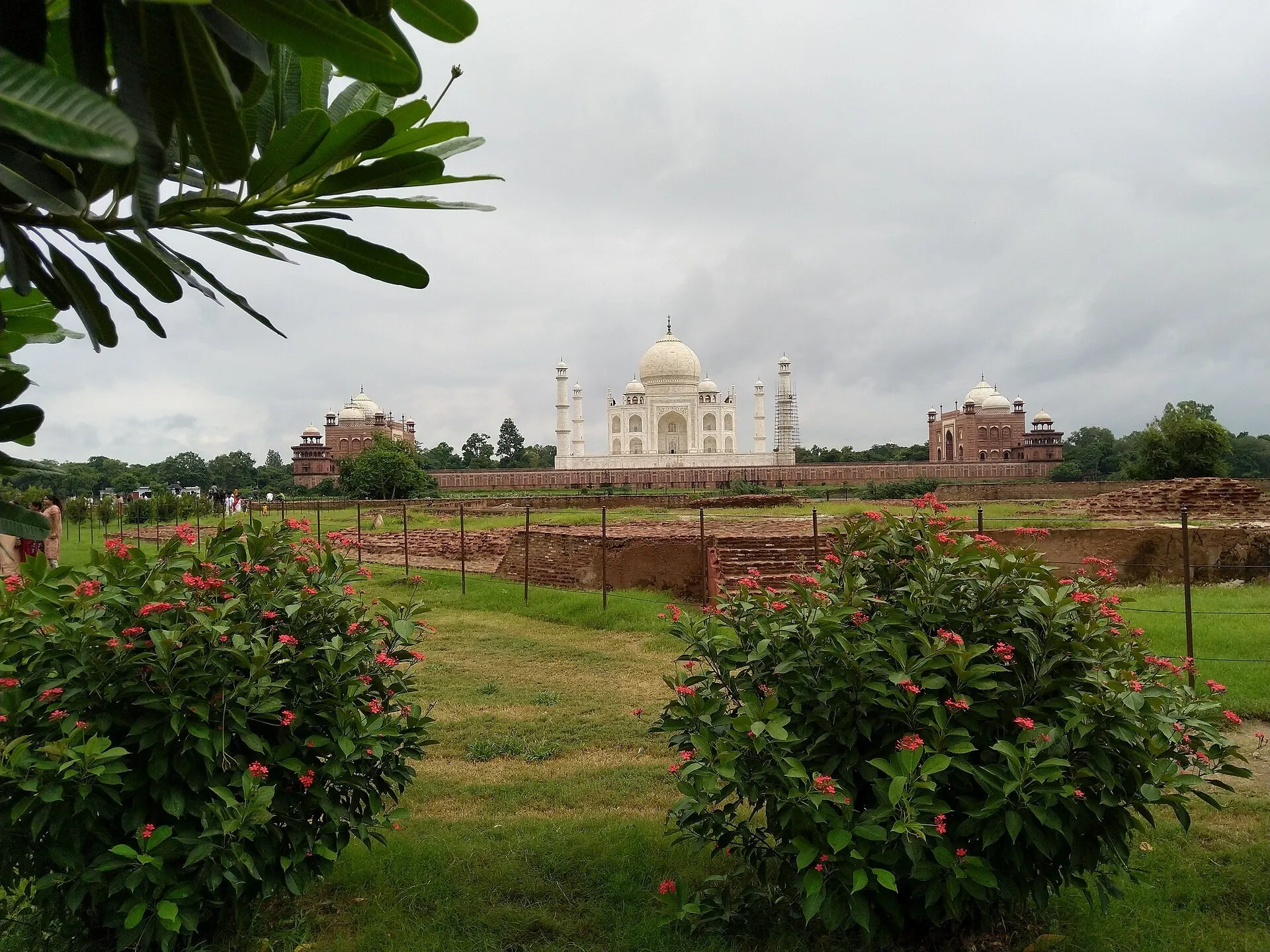 Jardín de Mehtab Bagh con flores y ruinas bajas; al fondo el Taj Mahal, con un minarete en restauración.