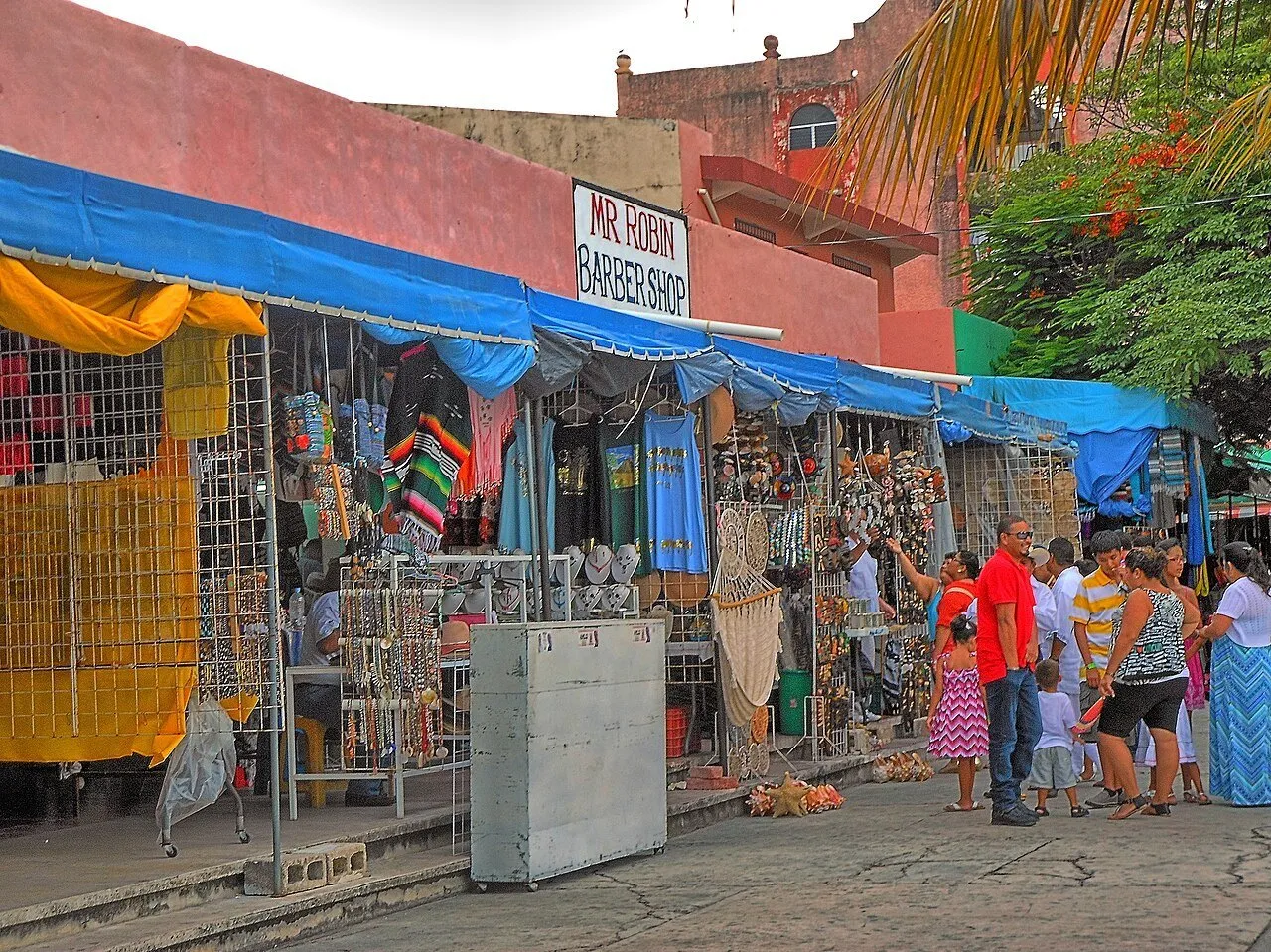 Lojas do Mercado 28 com toldos azuis, roupas e artesanato expostos e visitantes no corredor.