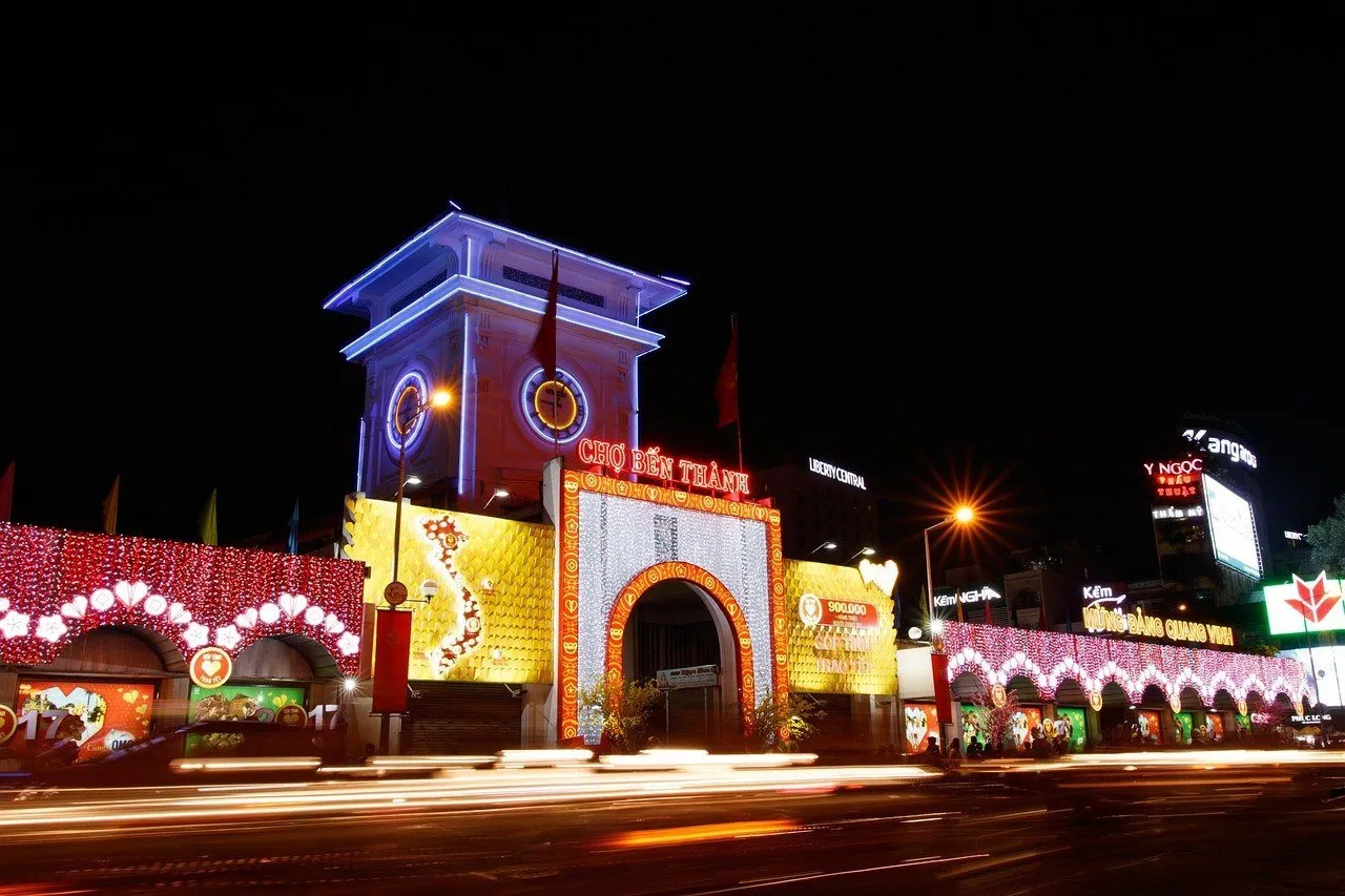 Entrada do Mercado Bến Thành iluminada à noite, com torre do relógio e rastros de luz dos veículos.