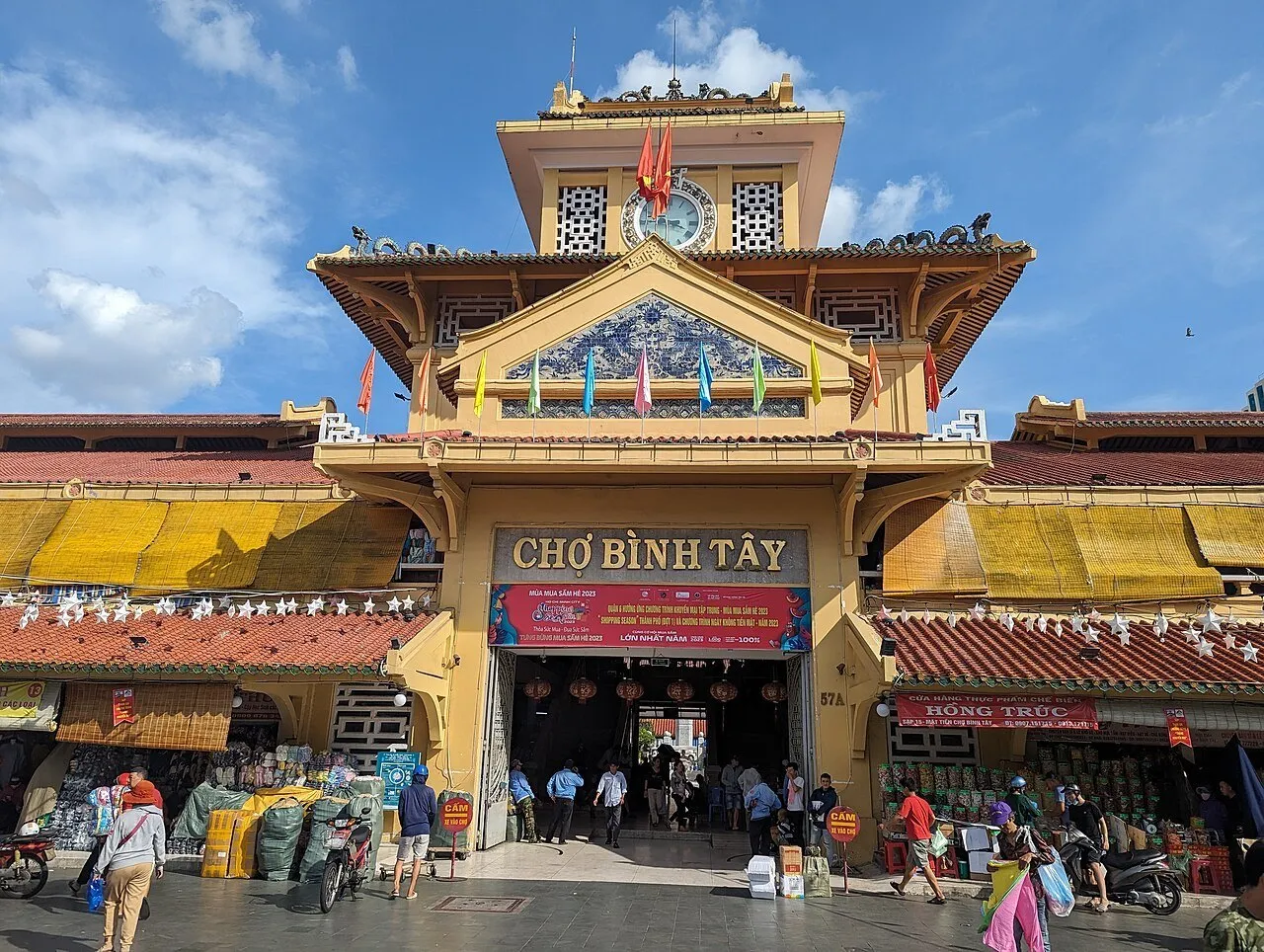 Entrada amarela do Mercado Bình Tây com torre do relógio, telhados decorados e comércio junto às portas.