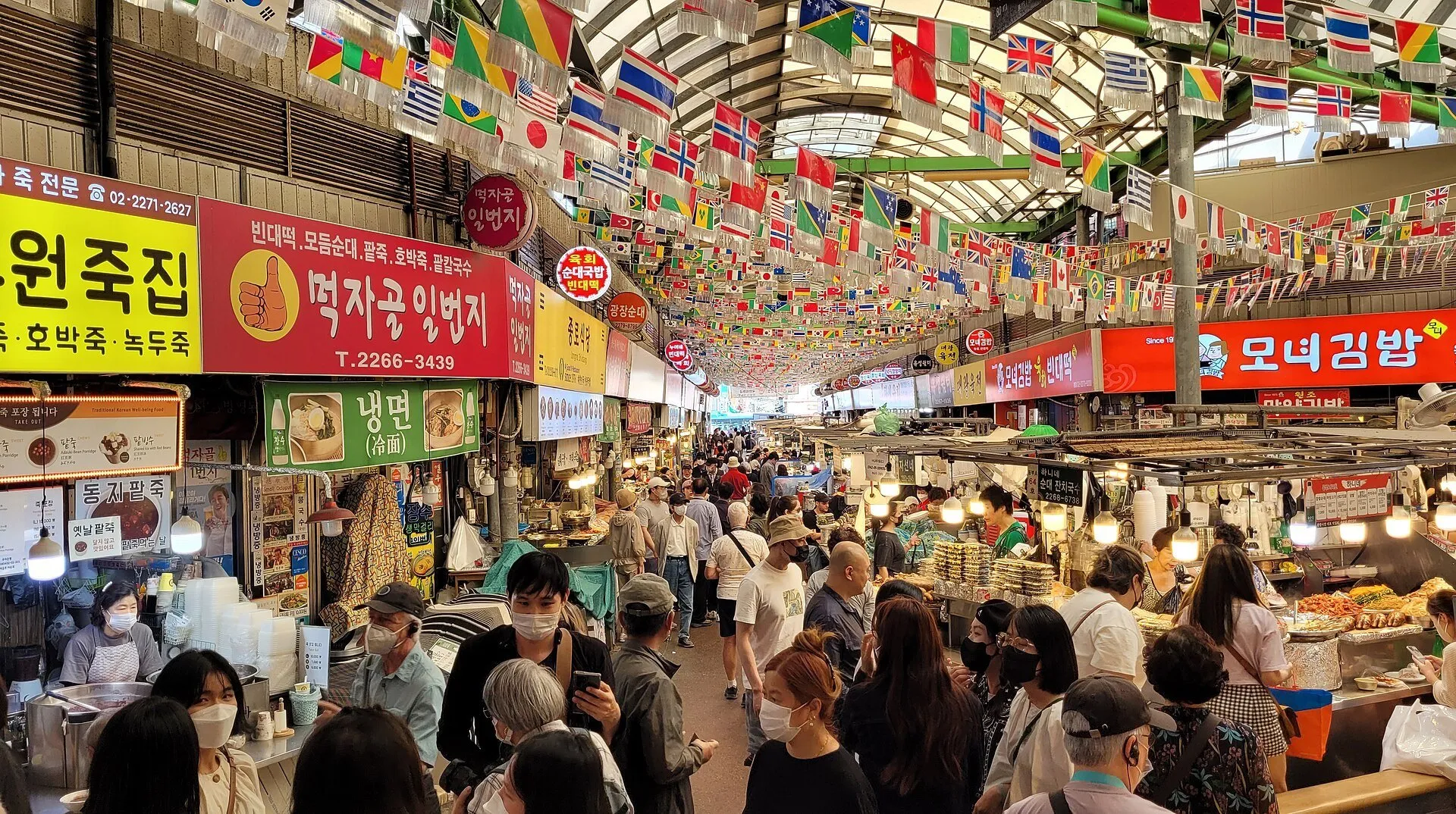 Pasillo cubierto del Mercado Gwangjang con letreros, banderas, puestos de comida y visitantes.