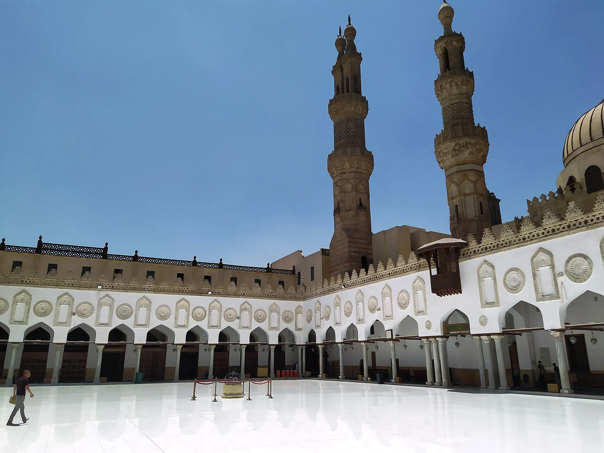 Pátio branco da Mesquita Al-Azhar no Cairo, cercado por arcadas, com dois minaretes e poucos visitantes.