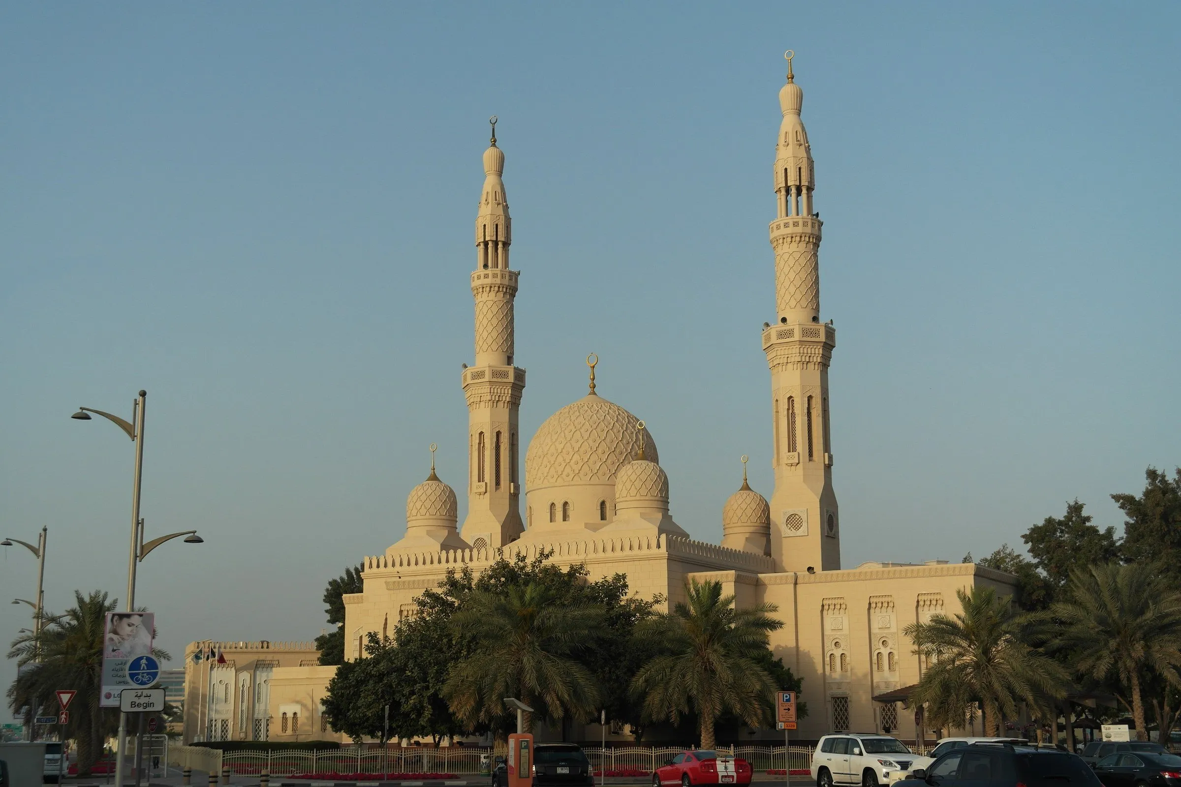 Full view of Jumeirah Mosque, with two minarets and pale stone domes among palm trees, in golden late-afternoon light.