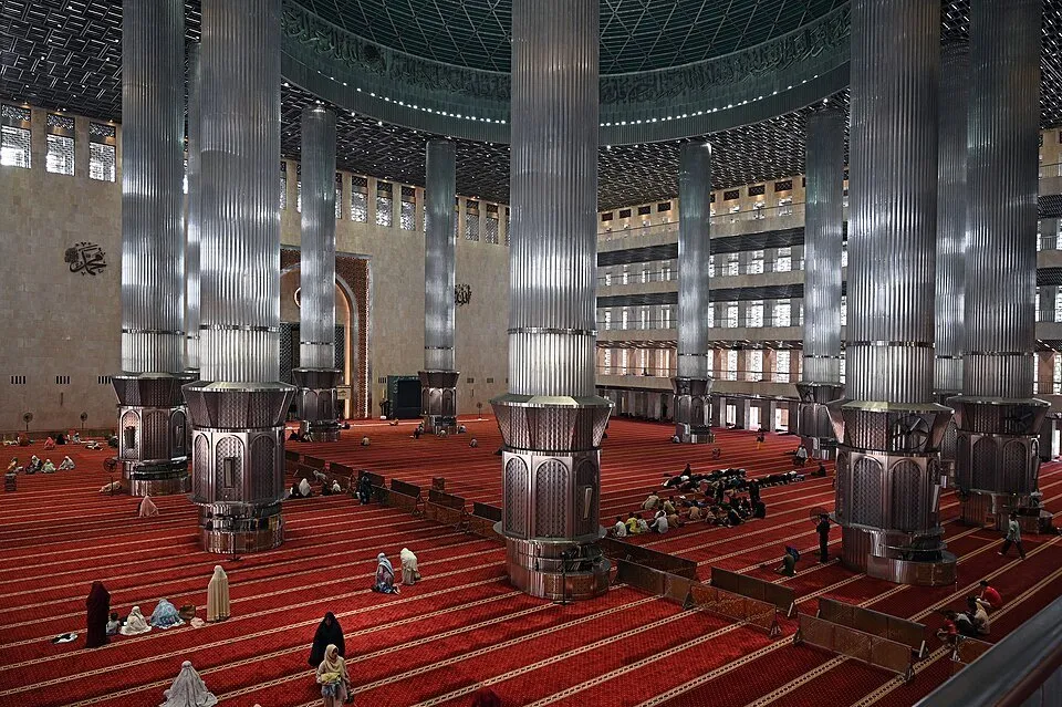 Salão da Istiqlal com grandes colunas metálicas, tapete vermelho e visitantes vistos da galeria.