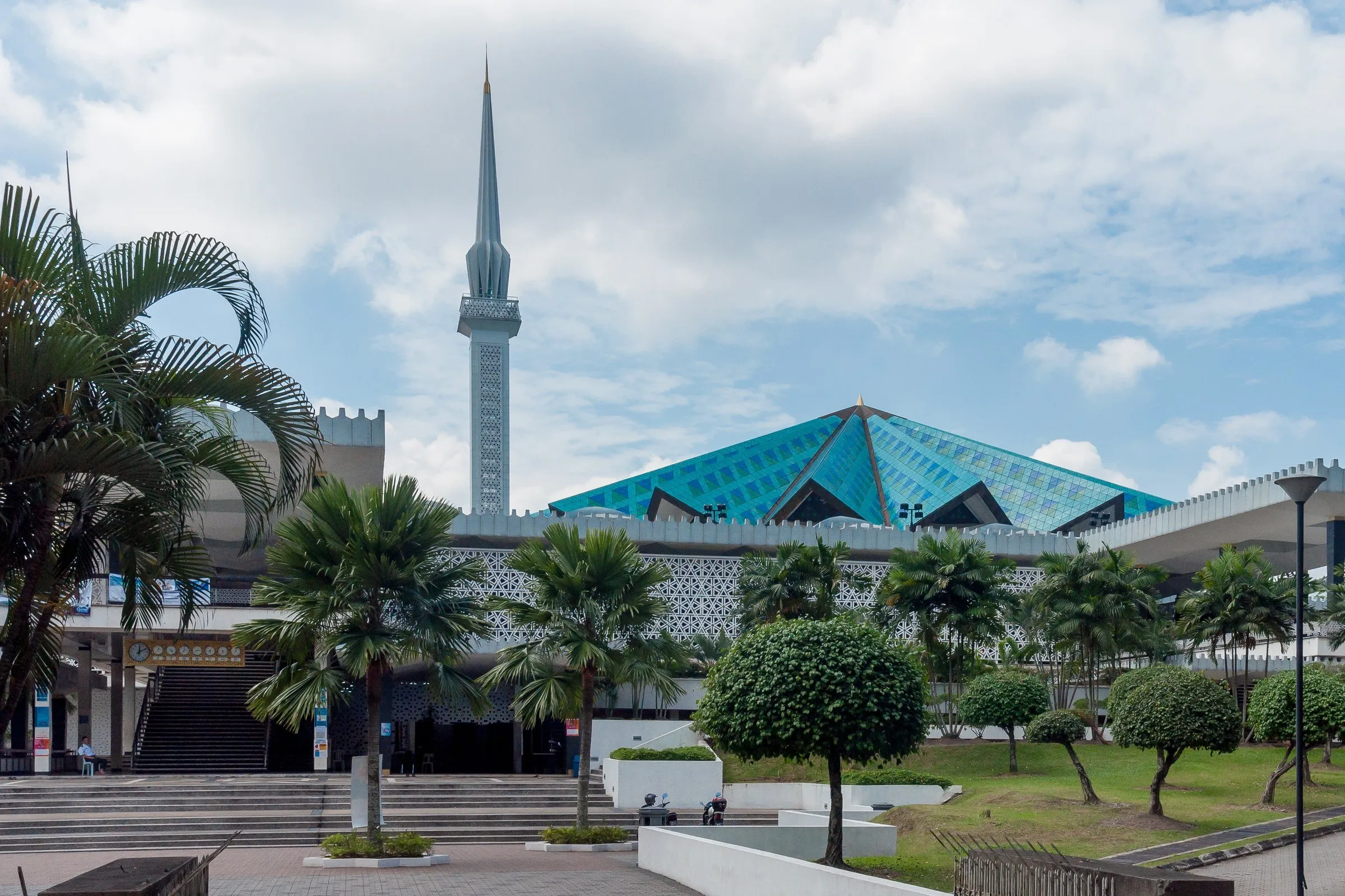 Telhado dobrado azul-turquesa e minarete alto da Mesquita Nacional atrás de palmeiras e jardim, sob céu com nuvens.
