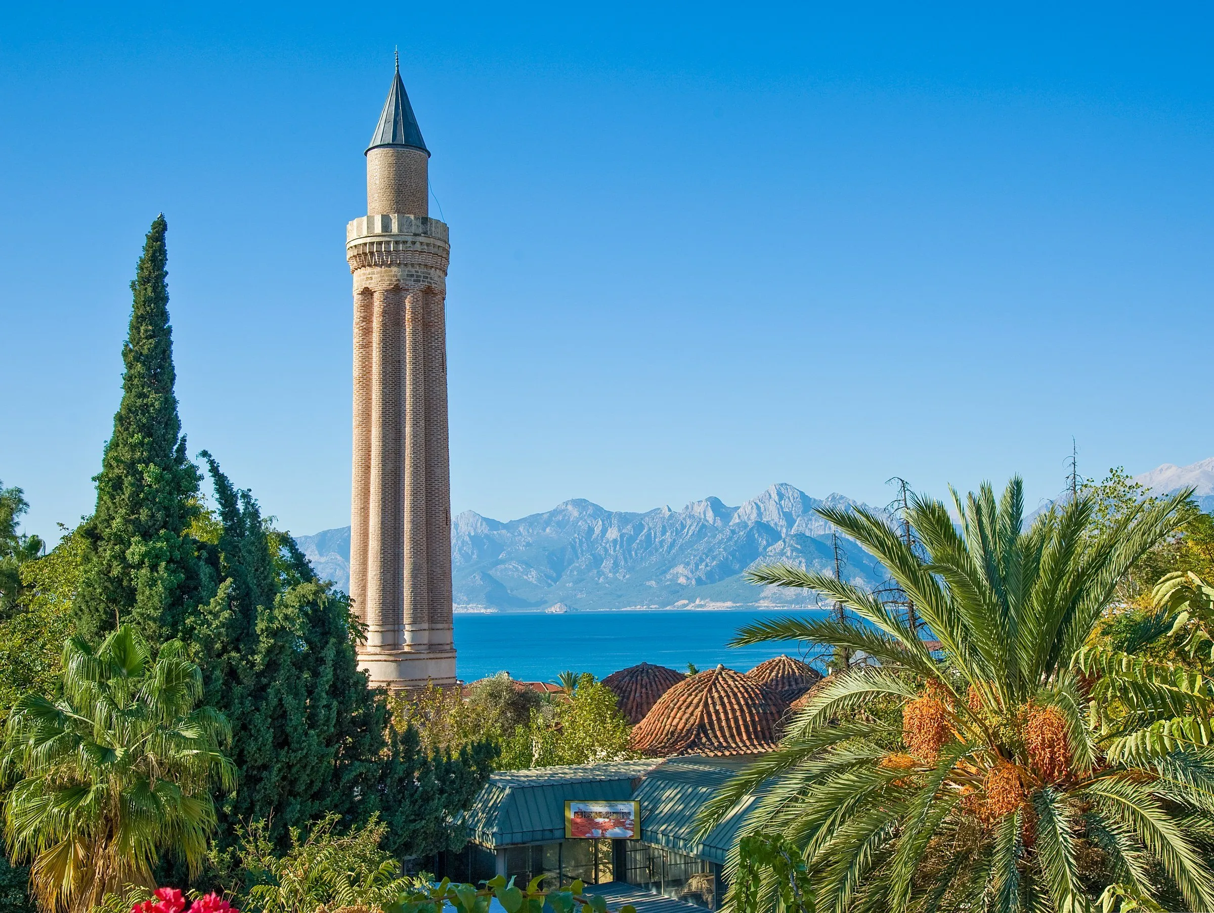 Minarete de tijolo com caneluras e ponta cônica entre palmeiras e ciprestes, com o mar e montanhas ao fundo sob céu azul.