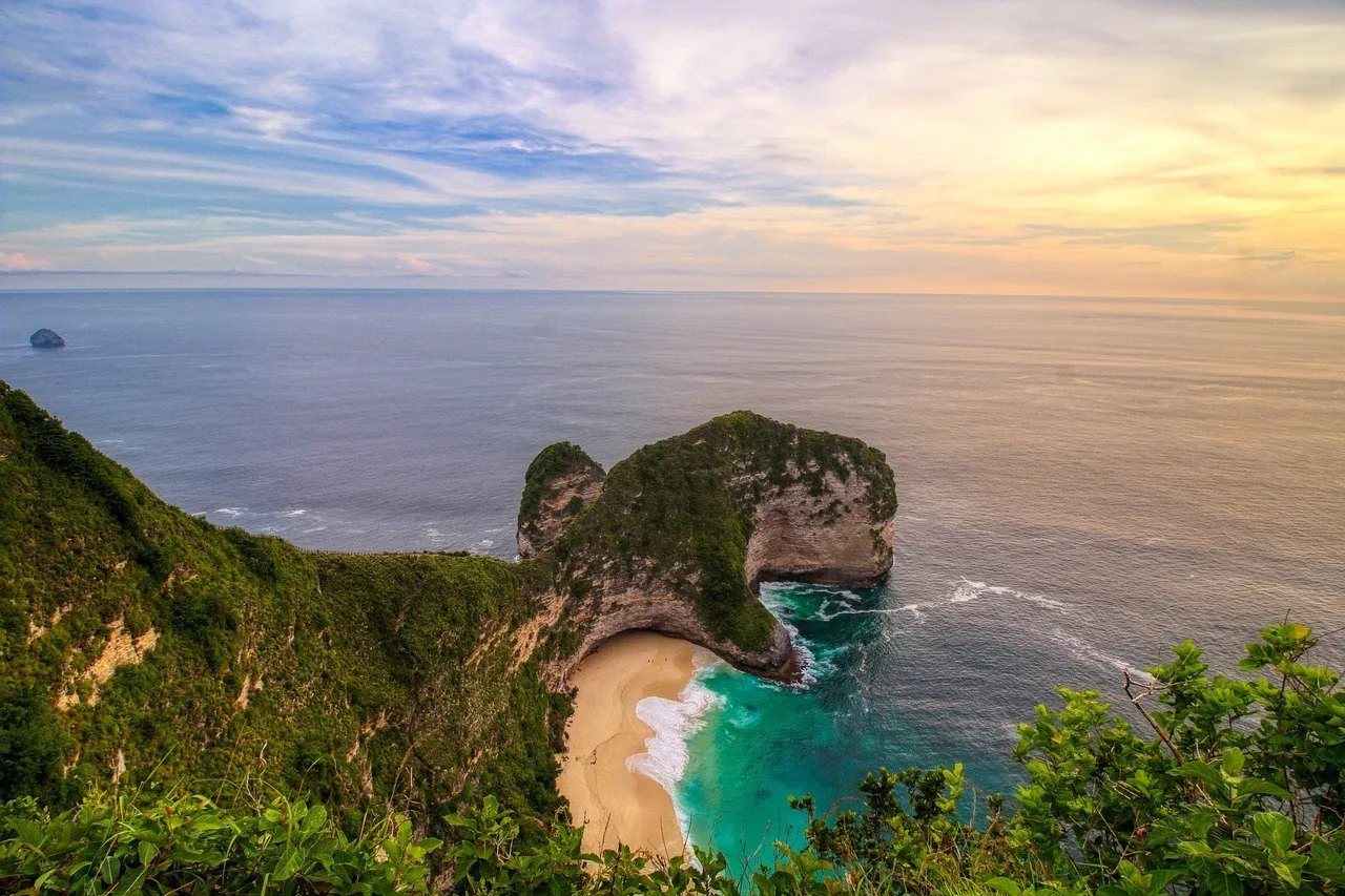 Acantilado de Kelingking visto desde arriba al atardecer, con playa clara, vegetación y mar azul verdoso.