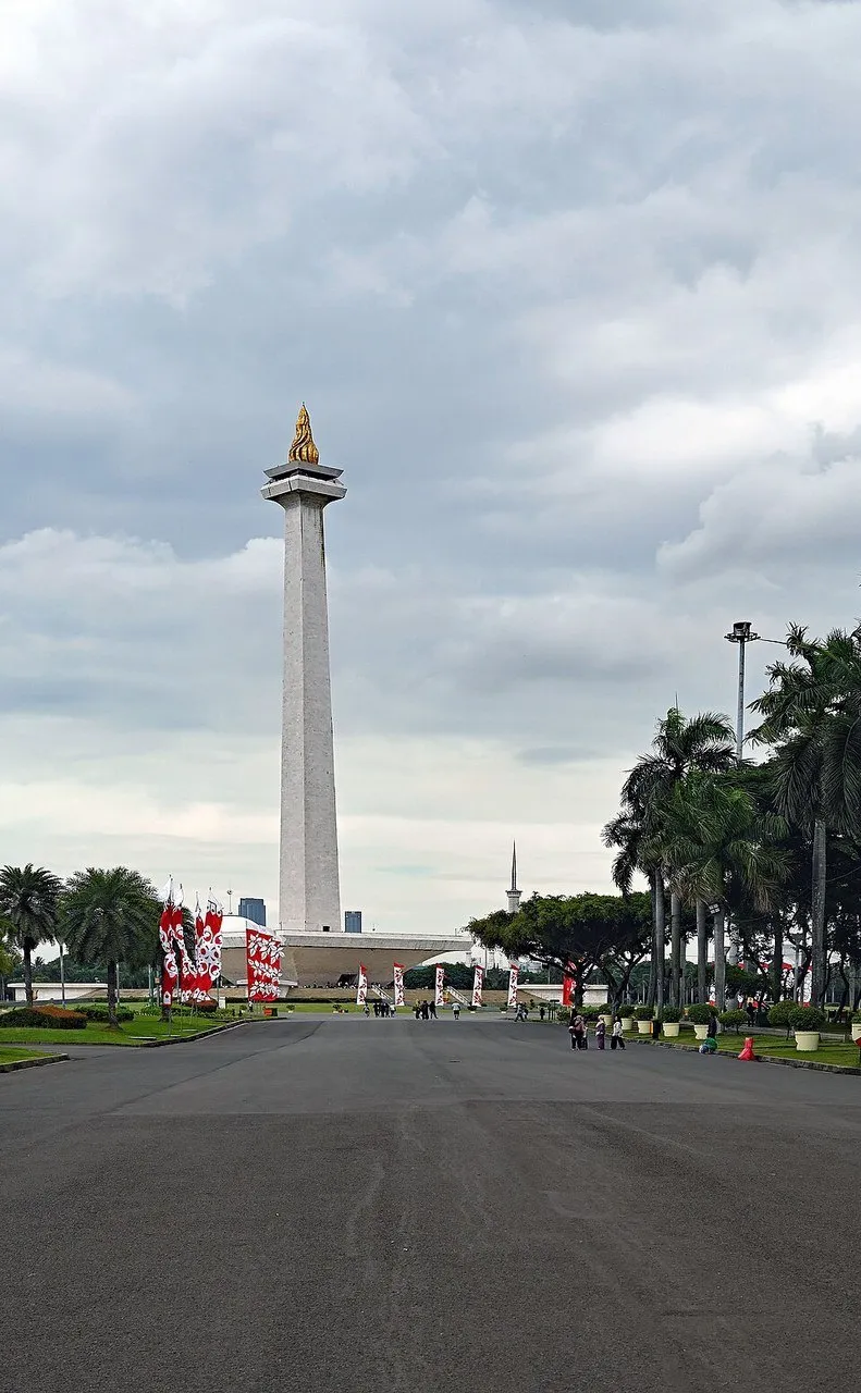 Monas inteiro com chama dourada, esplanada, palmeiras e pequenos visitantes sob céu nublado.