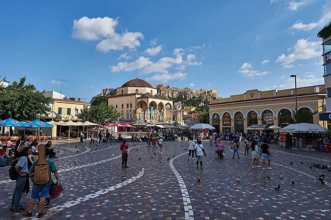 Monastiraki Square with pedestrians, a former mosque, a yellow station and the Acropolis behind under blue sky.