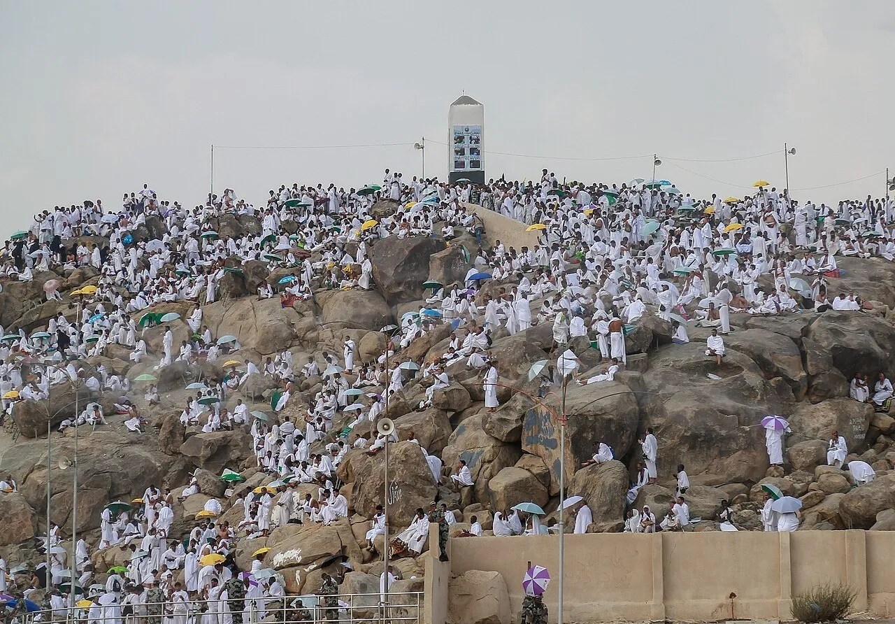 Encosta rochosa do Jabal al-Rahmah com um marco branco no topo e peregrinos espalhados entre as pedras.