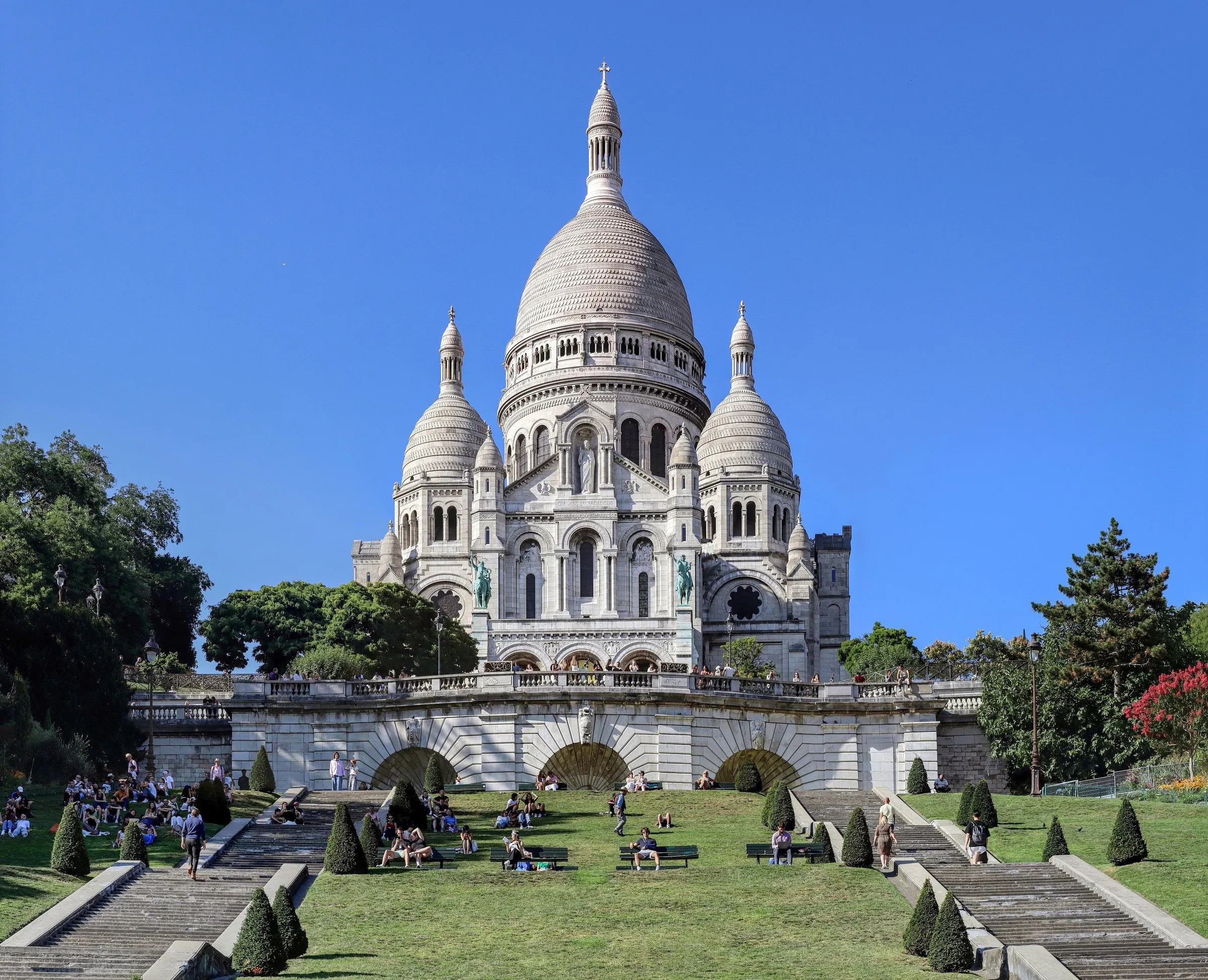 Basílica do Sacré-Cœur, de pedra branca e cúpulas, vista dos jardins em degraus de Montmartre, com gente sentada no gramado.