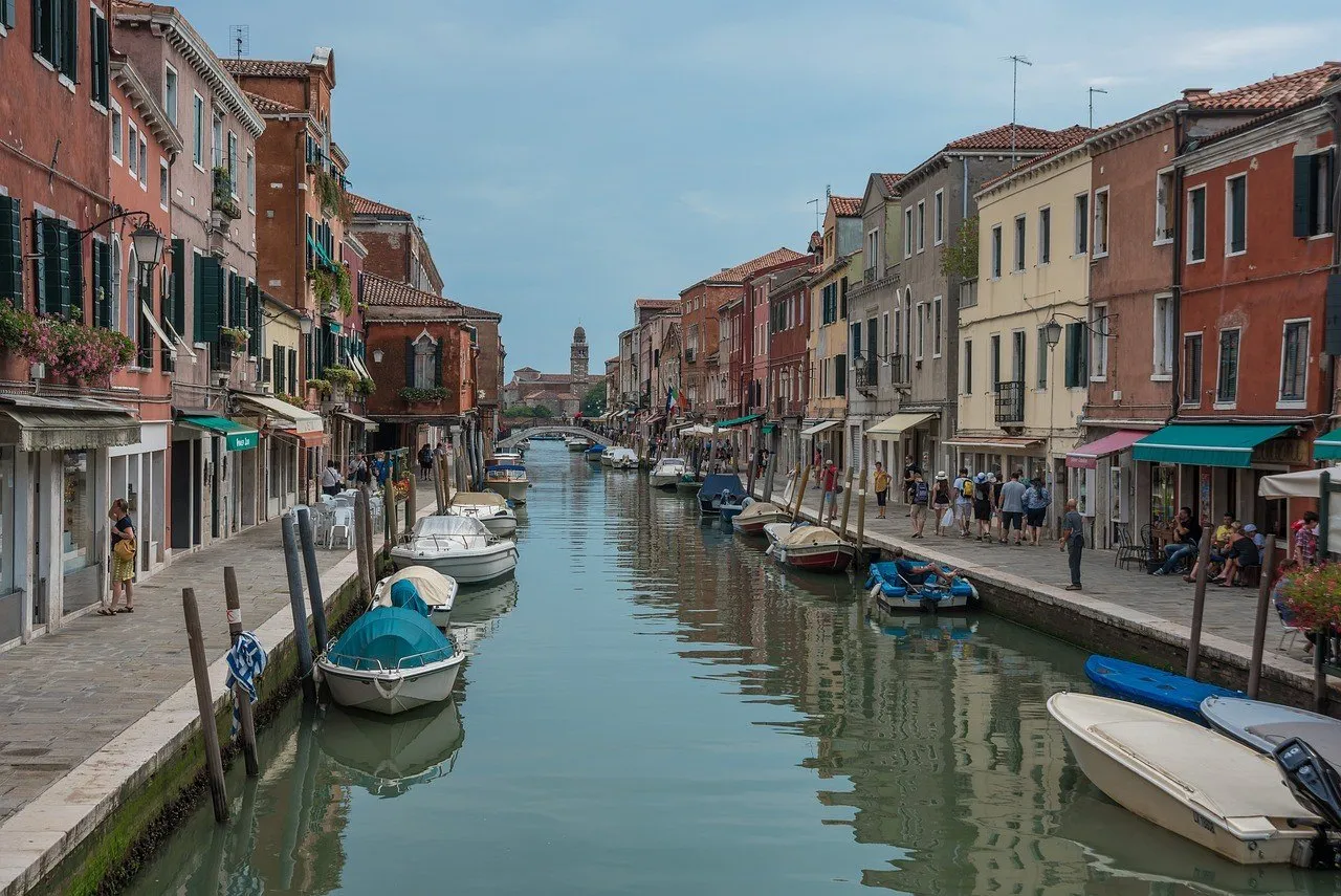 Canal de Murano ladeado por fachadas, com barcos amarrados, pedestres e uma ponte ao fundo.