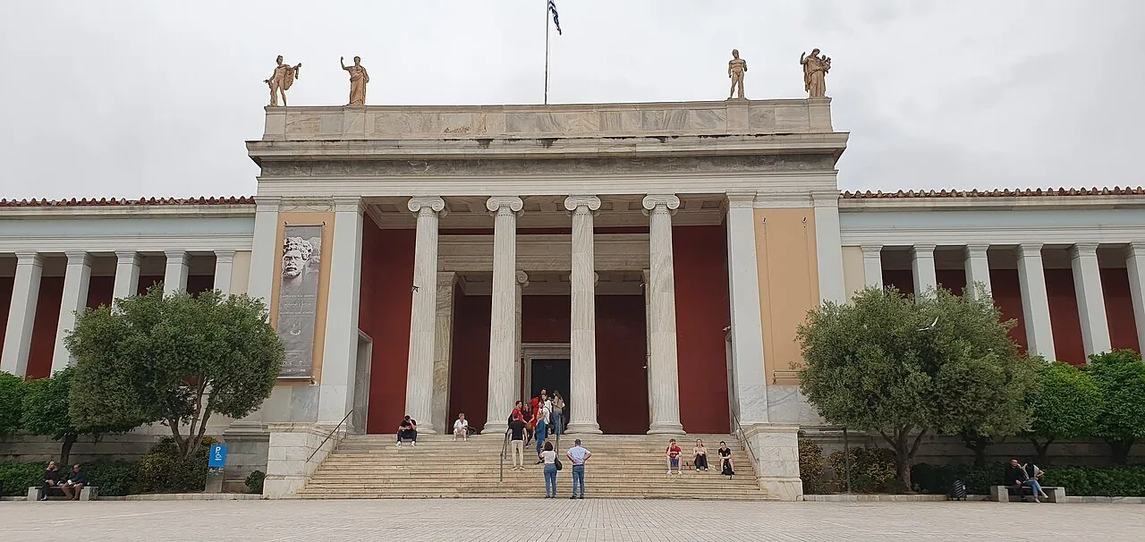 Facade of the National Archaeological Museum of Athens, with columns, a staircase and side trees under cloudy sky.