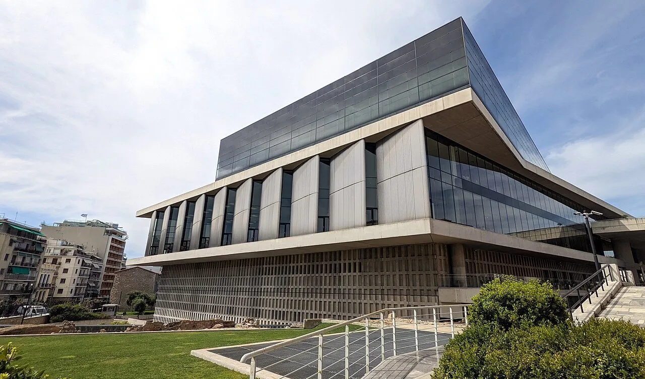 Exterior of the Acropolis Museum in Athens, with glass and concrete facades, a lawn and a ramp in front.