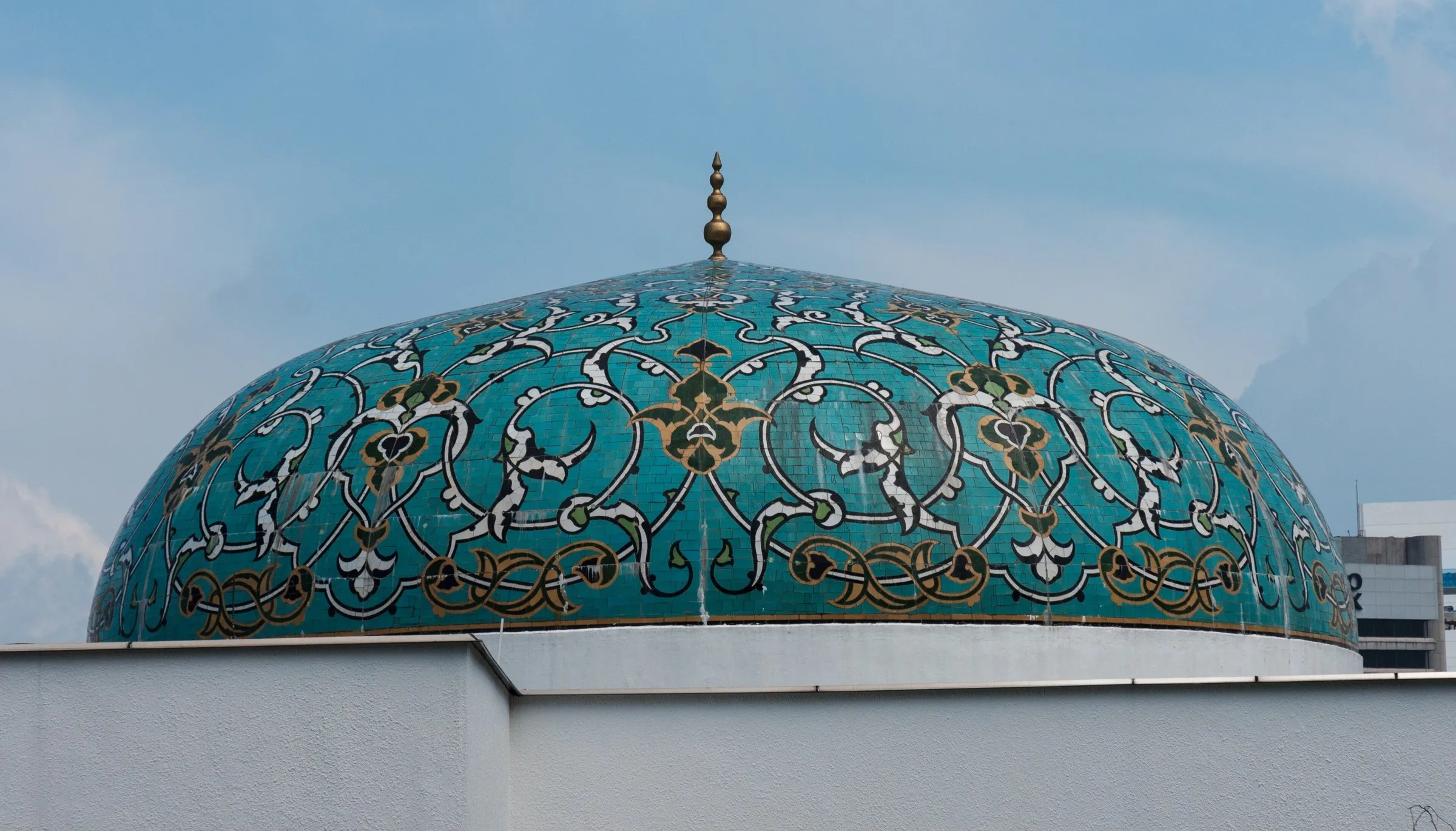 Cúpula de azulejos turquesa com arabescos florais em branco, verde e dourado, sob céu azul, com pináculo metálico no topo.