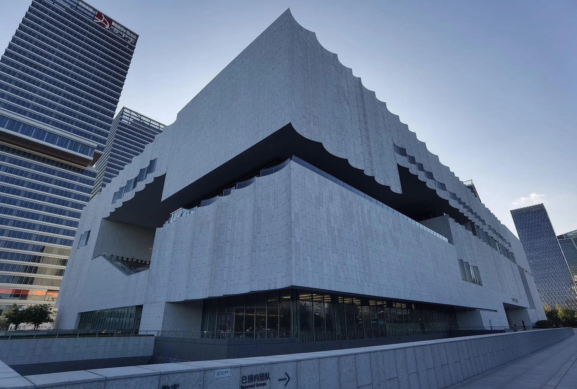 Light facade of the Shanghai Museum East, with wavy cutouts, windows and tall buildings around.