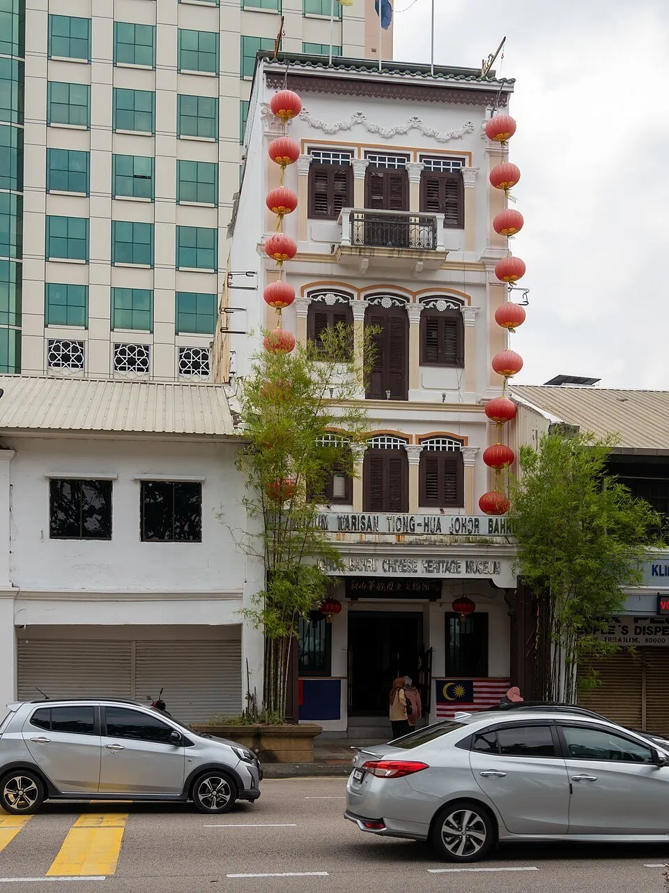Fachada estreita do Museu da Herança Chinesa de Johor Bahru, com lanternas vermelhas e carros na rua.