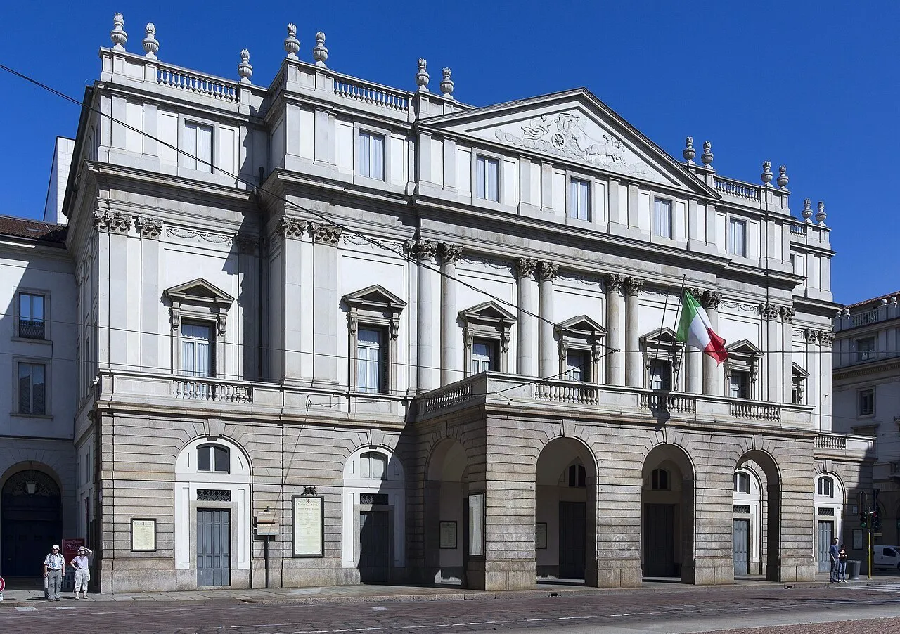 Light-colored façade of the Teatro alla Scala with portico, Italian flag and few pedestrians under a blue sky.