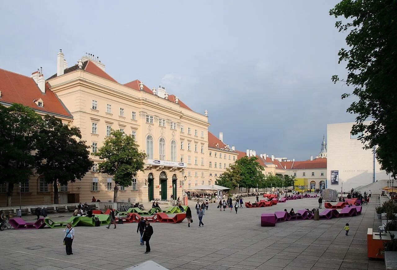 Pátio do MuseumsQuartier com bancos coloridos, visitantes e fachada clara do Leopold Museum à direita.