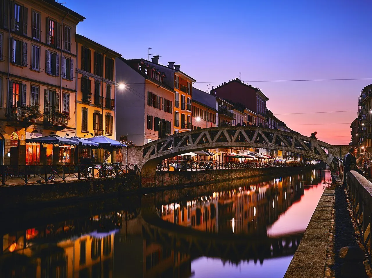 Alda Merini Bridge over the Naviglio Grande, with lit façades reflected in the water at dusk.