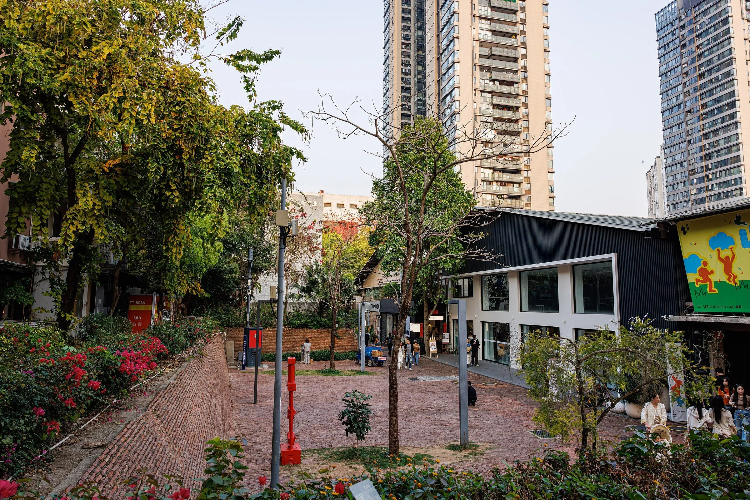Pracinha de tijolos no OCT-LOFT, com árvores, galpões reformados e torres residenciais ao fundo.
