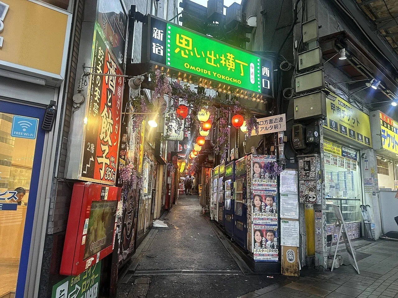 Entrada de Omoide Yokocho con letrero verde, faroles encendidos, pequeñas fachadas y pasillo estrecho.