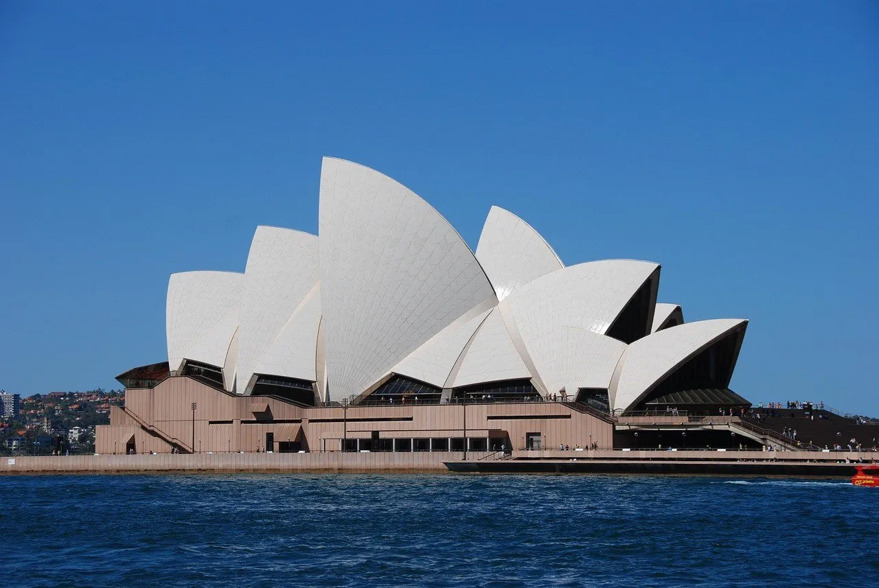 Sydney Opera House seen from the water, with white roofs, a stone base and blue sky.