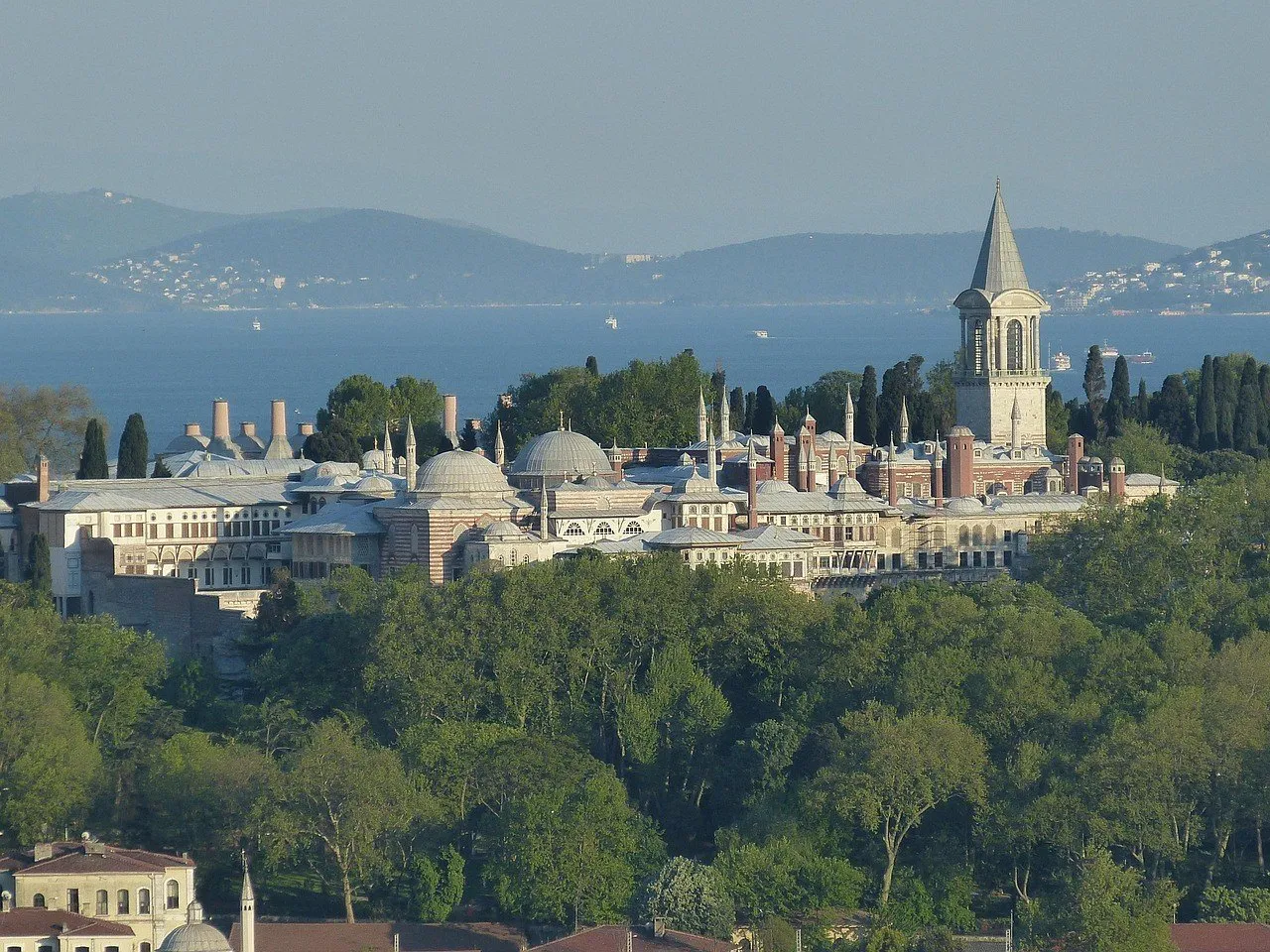 Conjunto del Palacio de Topkapi entre árboles, con la Torre de la Justicia de techo cónico a la derecha y el mar al fondo.
