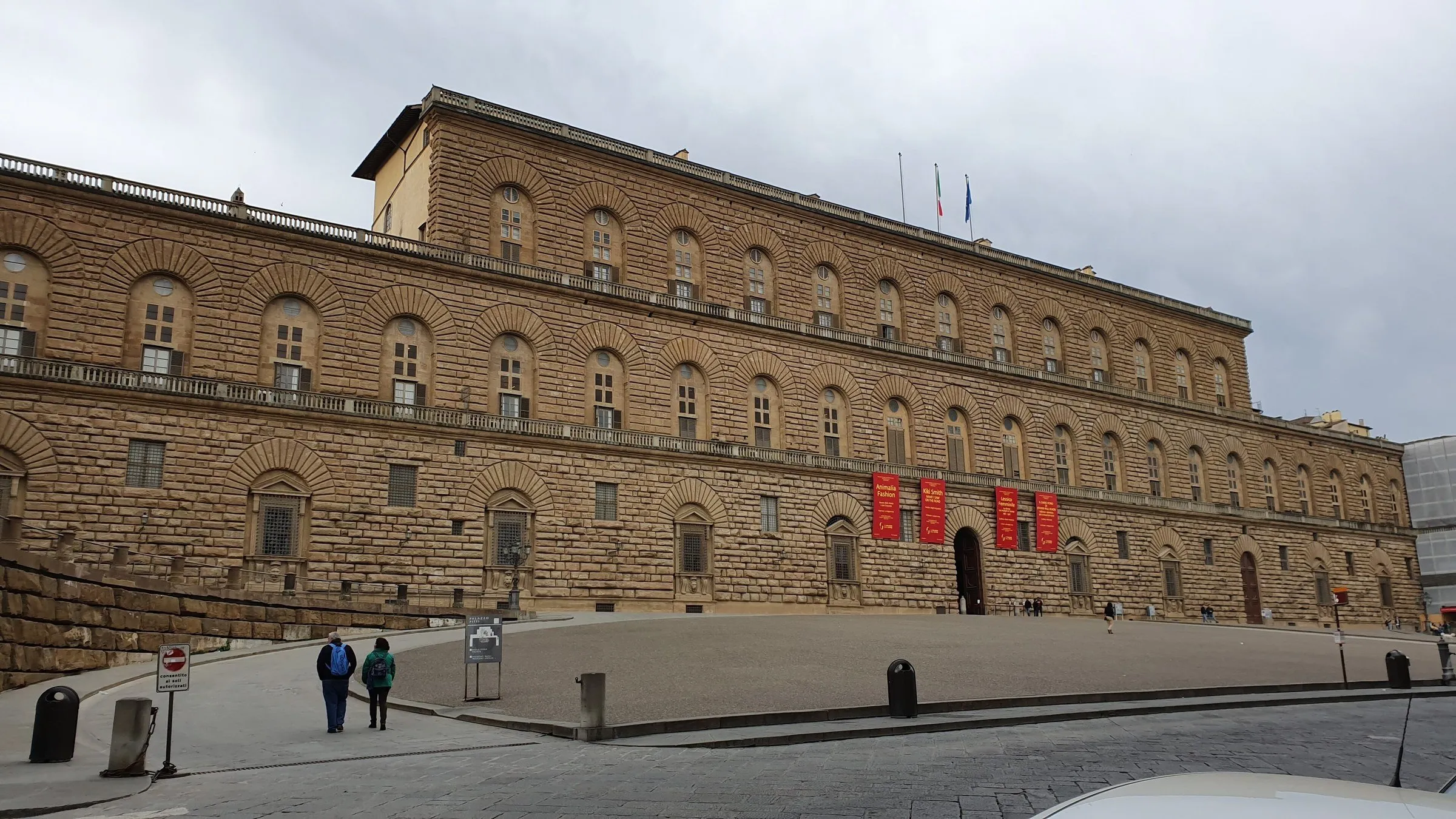 Stone facade of Palazzo Pitti and sloping square, with few people and an overcast sky.