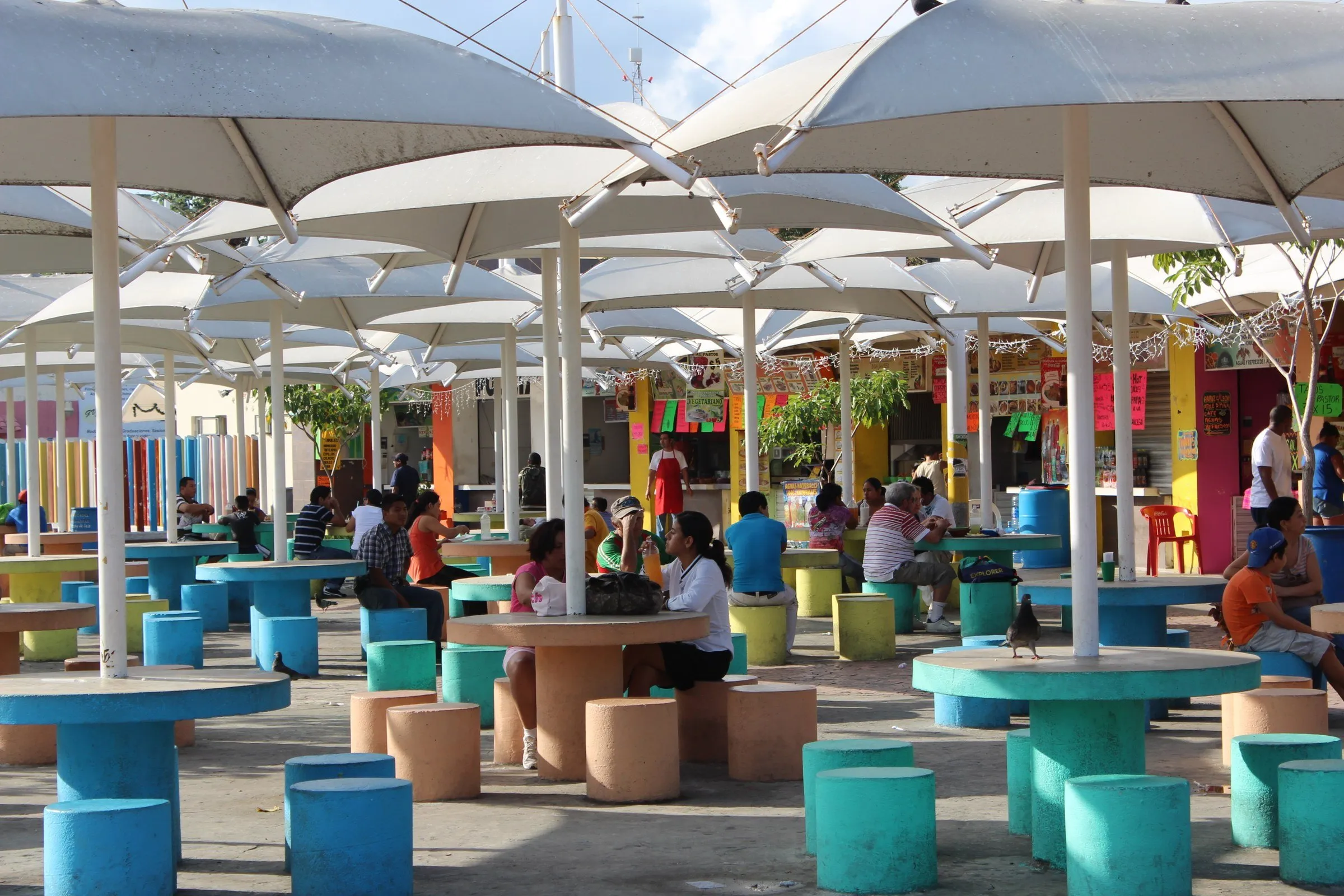 Mesas e bancos coloridos sob coberturas brancas na área de alimentação do Parque de las Palapas, com pessoas ao fundo.