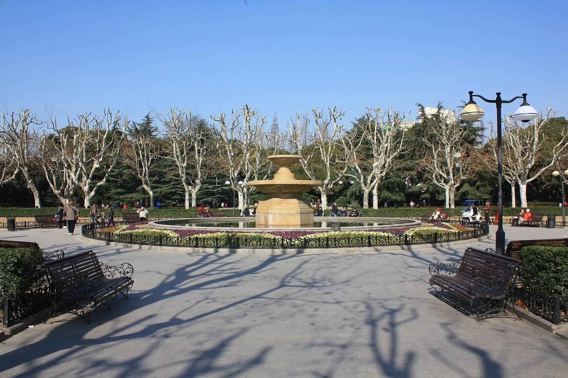 Circular fountain among flower beds, benches and leafless trees in Fuxing Park, under a blue sky.