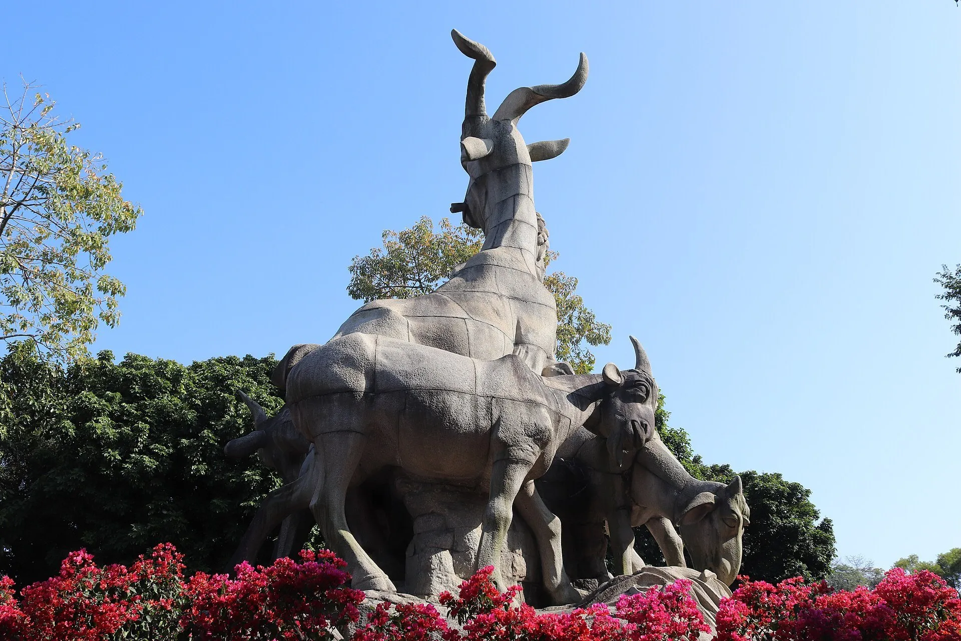 Escultura de las cabras vista de lado en el Parque Yuexiu, entre flores rojas, árboles y cielo azul.