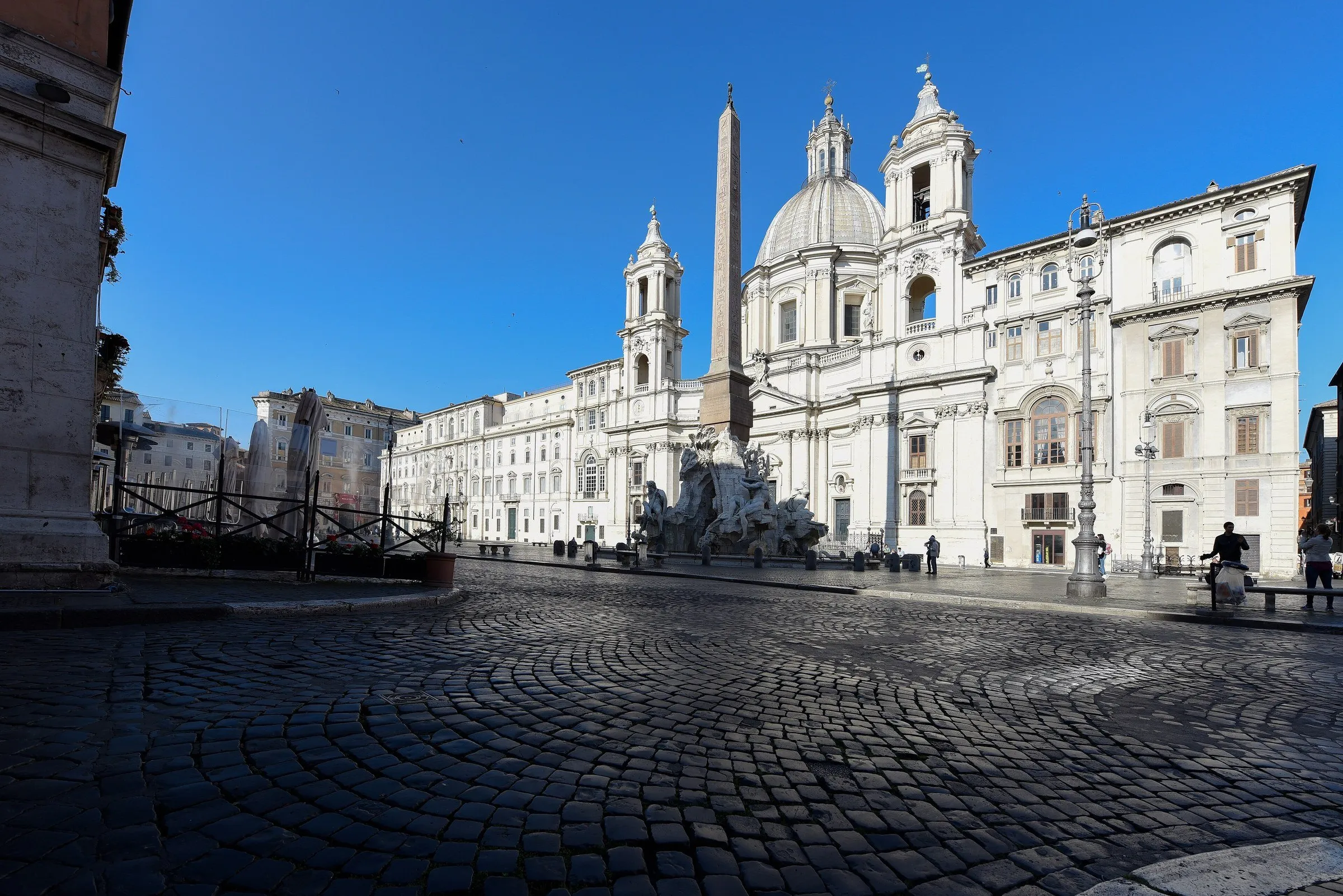 Cobblestone square with the church of Sant'Agnese in Agone, the obelisk and the Fountain of the Four Rivers under a blue sky.