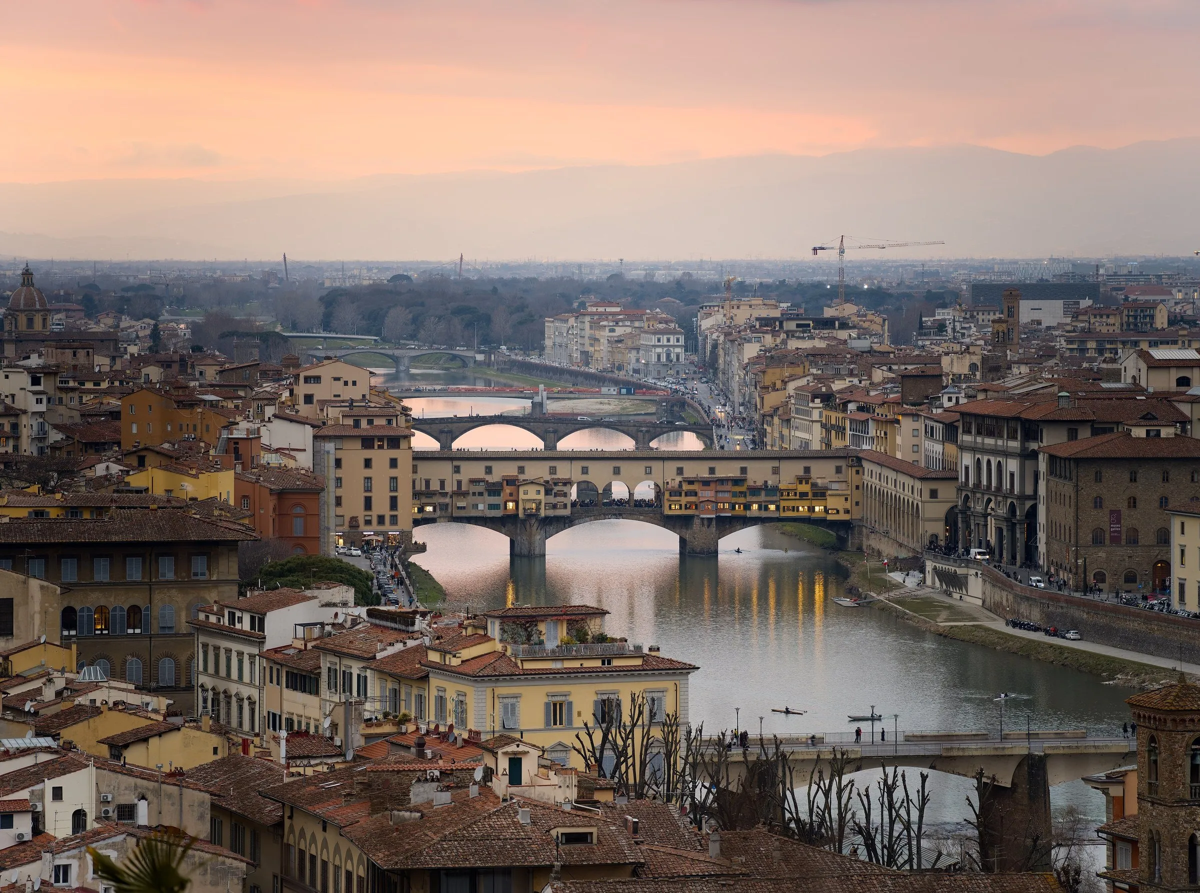Elevated view of the Ponte Vecchio and other bridges over the Arno, with rooftops and a pink sky.