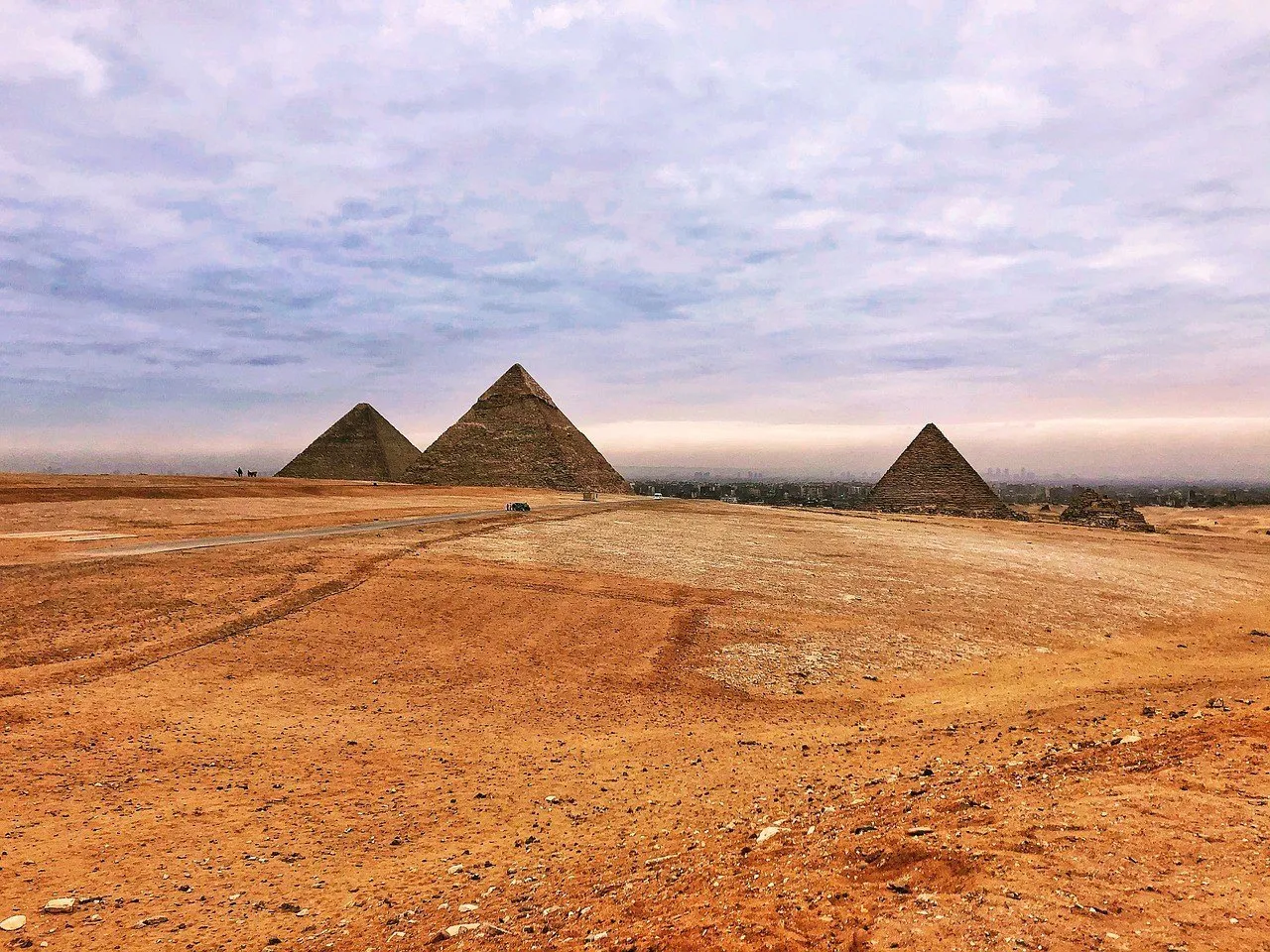 Panorama das três pirâmides de Gizé, com chão desértico em primeiro plano e céu de nuvens rosadas.
