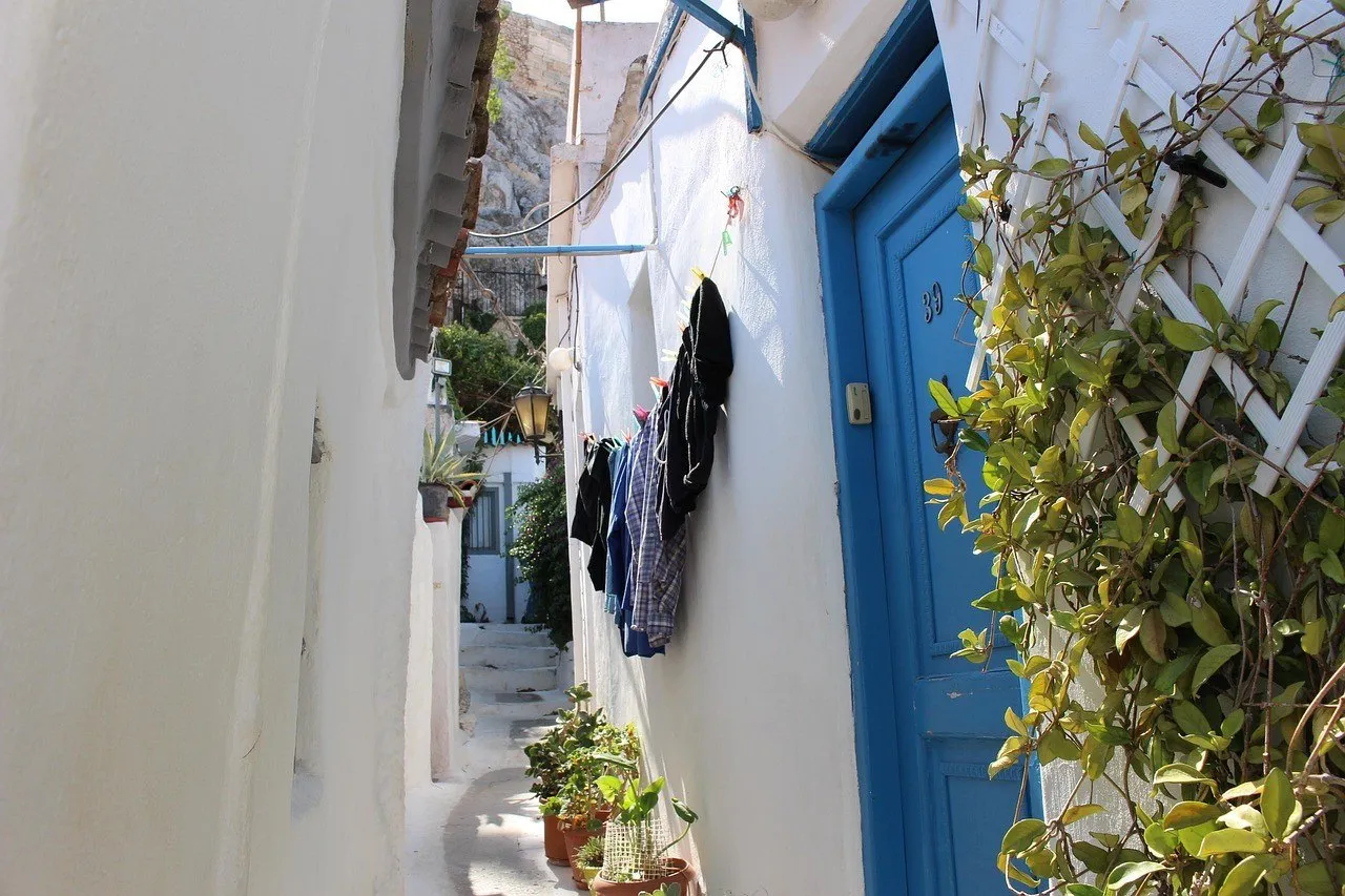 Narrow passage in Plaka, Athens, with white walls, a blue door, plants and steps.