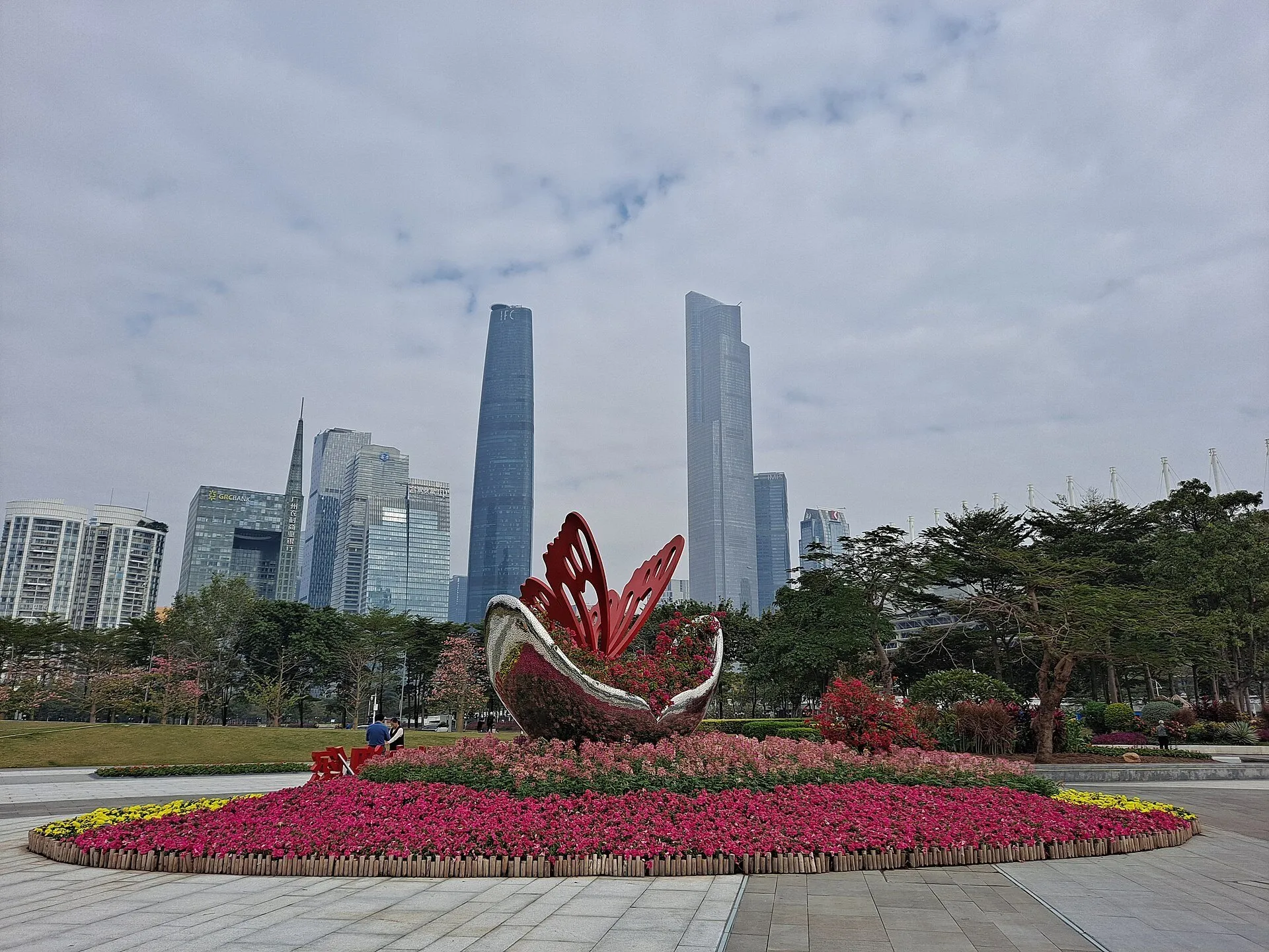 Parterre de flores con escultura roja en la Plaza Huacheng, frente a rascacielos bajo un cielo nublado.