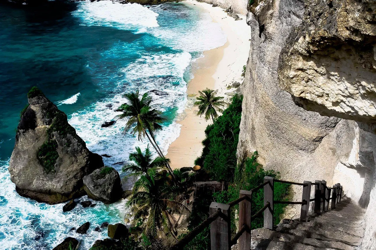 Escalera en el acantilado de Diamond Beach, con cocoteros, peñasco puntiagudo, arena clara y olas azules en Nusa Penida.