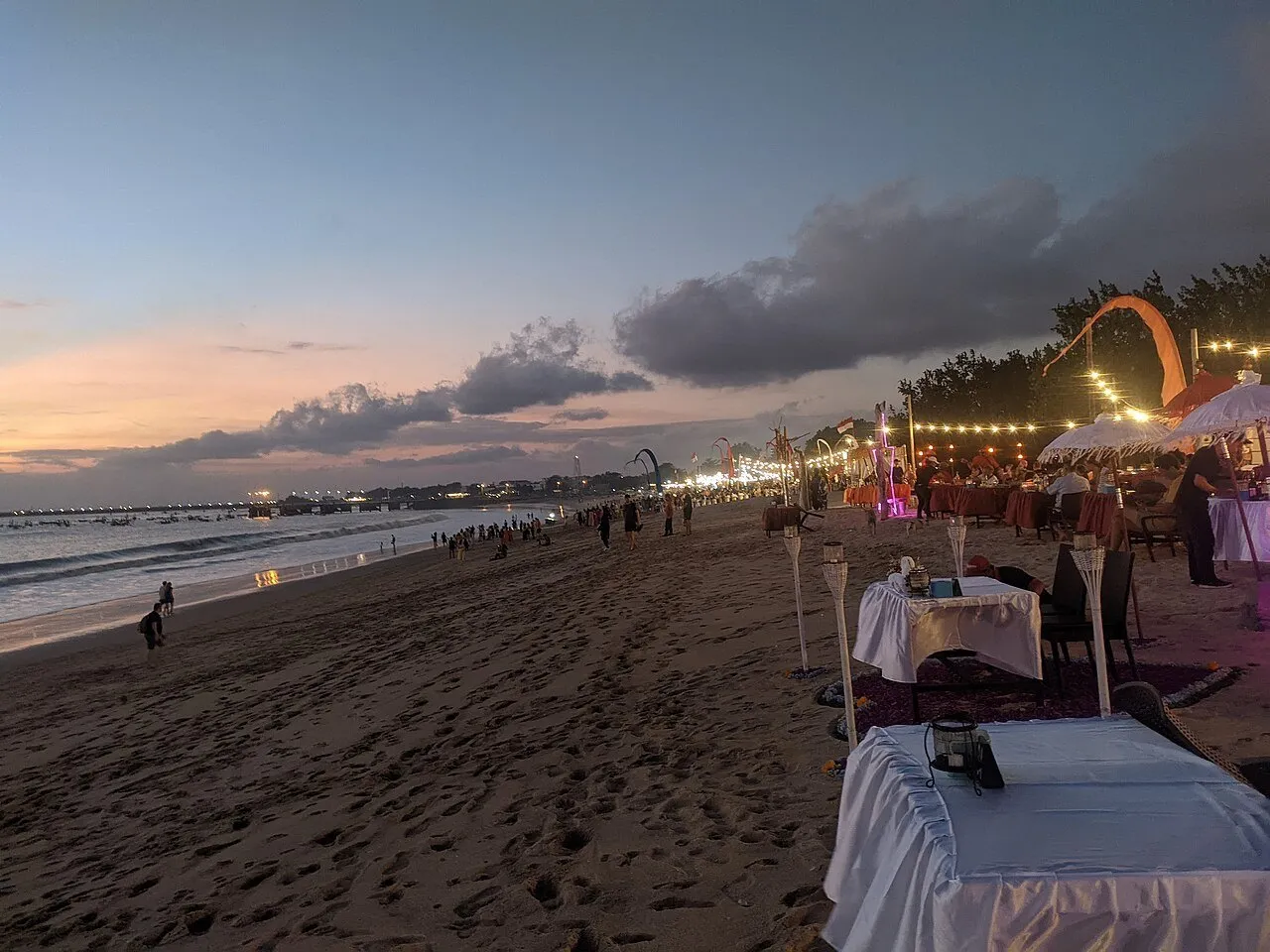 Mesas de restaurantes sobre la arena de Jimbaran al atardecer, con guirnaldas de luces, mar y nubes al fondo.