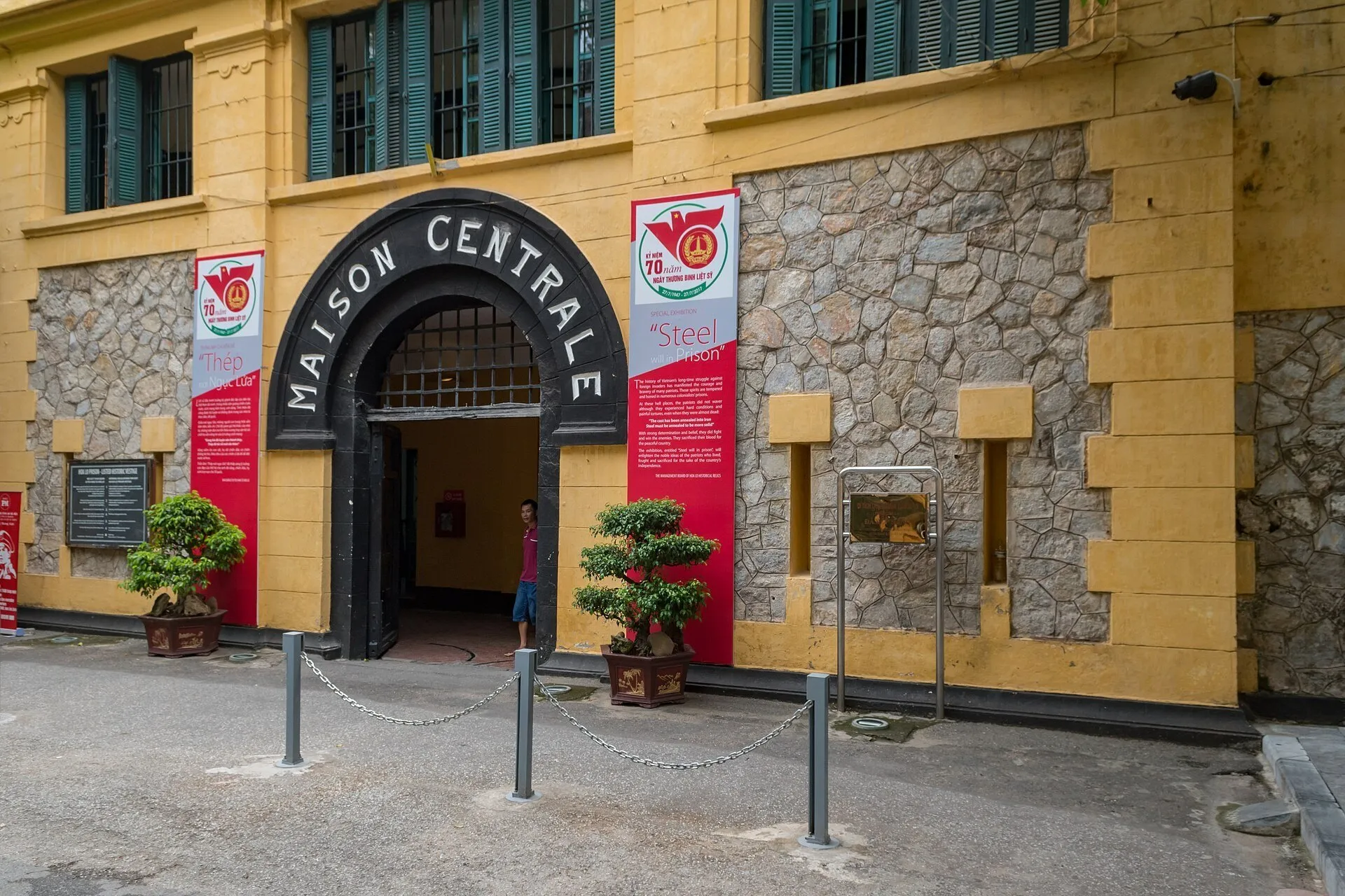 Entrada da Prisão Hoa Lo com arco Maison Centrale, fachada amarela, pedras e vasos de plantas.